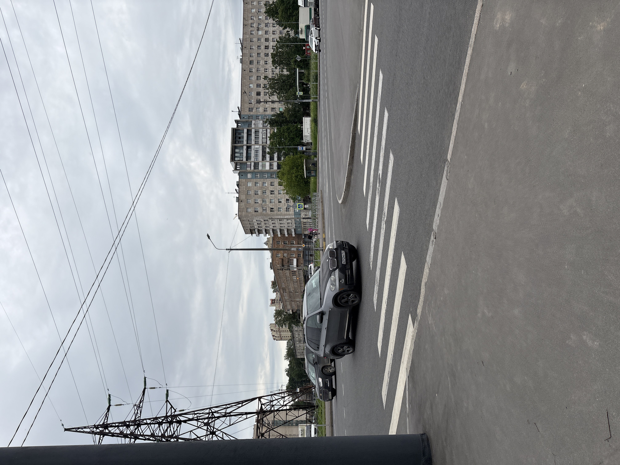 The image captures an intersection in Saint Petersburg, Russia, on a cloudy day. A large power line tower is prominently visible on the left, with thick cables stretching across the sky. The road features a white pedestrian crossing. In the foreground, a dark grey BMW X5 is stopped at the crossing, with its license plate reading "M312MX 178". To its left, another dark SUV, possibly a Kia Sorento, is also present. In the background, a row of multi-story apartment buildings lines the street, characteristic of Soviet-era architecture. Trees and a bus stop shelter are also visible. A white truck is turning at the intersection. The overall scene depicts a typical urban environment in Saint Petersburg.