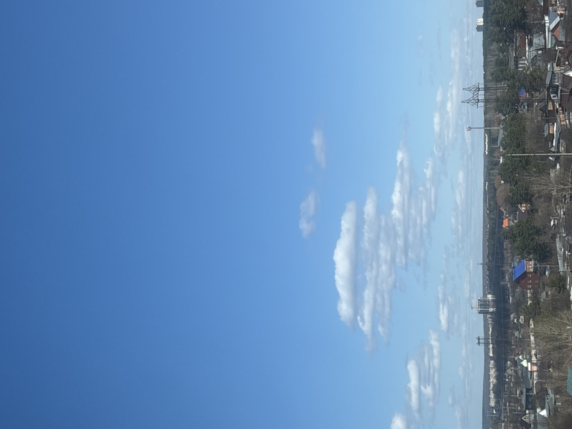 The image presents a bright blue sky with scattered elongated white clouds filling the right half of the frame. The horizon line features a dense urban landscape, likely Chaikovsky, Russia, characterized by a mix of residential buildings, trees, and infrastructure. Utility poles and power lines are visible amidst the buildings, suggesting an electrical distribution network. The architecture appears varied, with a mix of low-rise houses and taller structures. The overall impression is of a clear day in a moderately populated urban area.