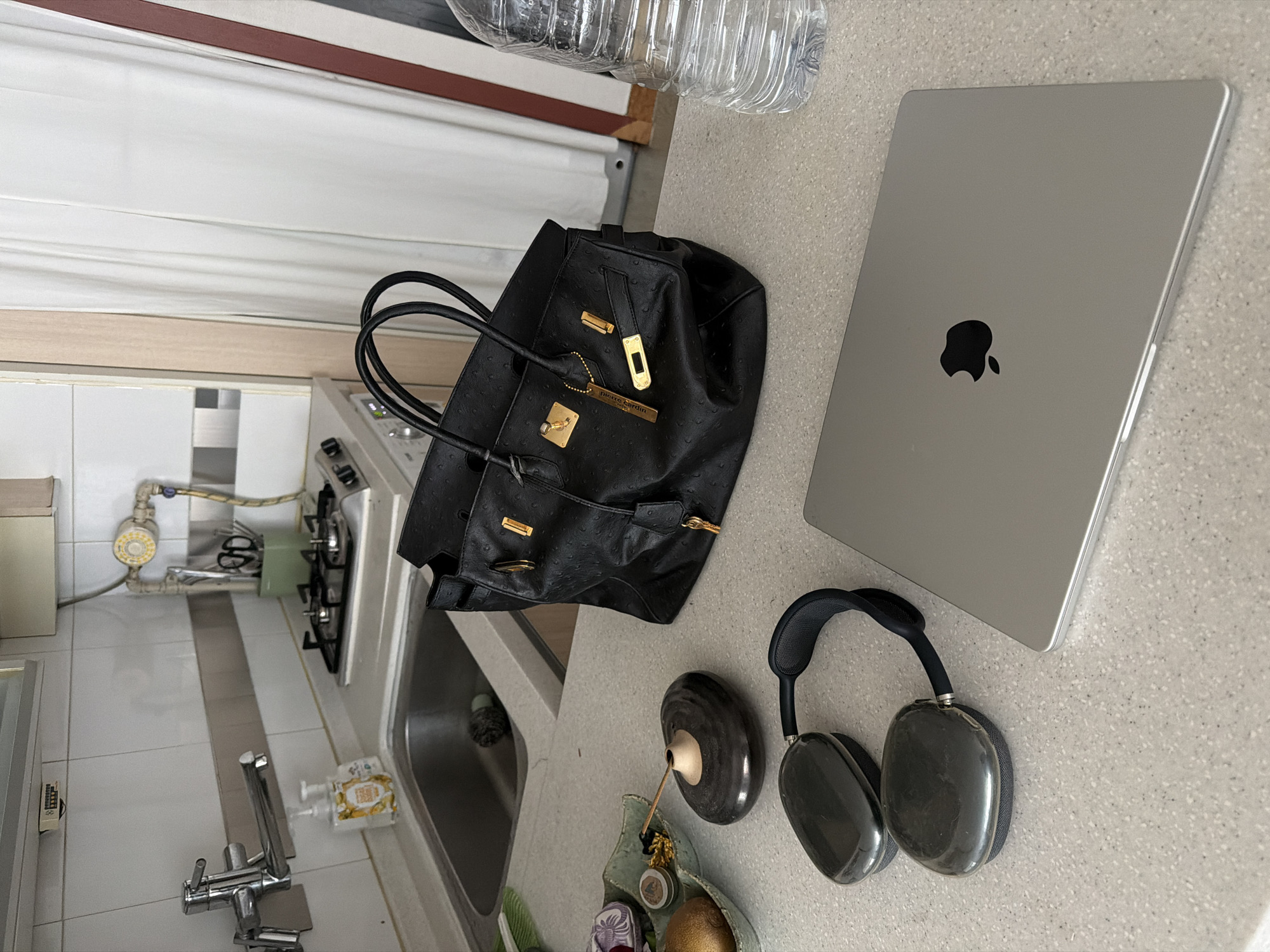 The image captures a scene in a domestic kitchen in Seoul, South Korea. A gray countertop is centrally positioned and supports several items: a closed silver-colored MacBook, a black leather handbag with gold hardware, a pair of dark gray headphones, a bottle of water, an incense holder with a lit incense stick, and a decorative dish holding fruits and small objects. The kitchen area is visible to the left, featuring a sink, a gas stove, and typical kitchen appliances and fixtures. The color palette is dominated by gray, black, and metallic tones, creating a modern and minimalist aesthetic.