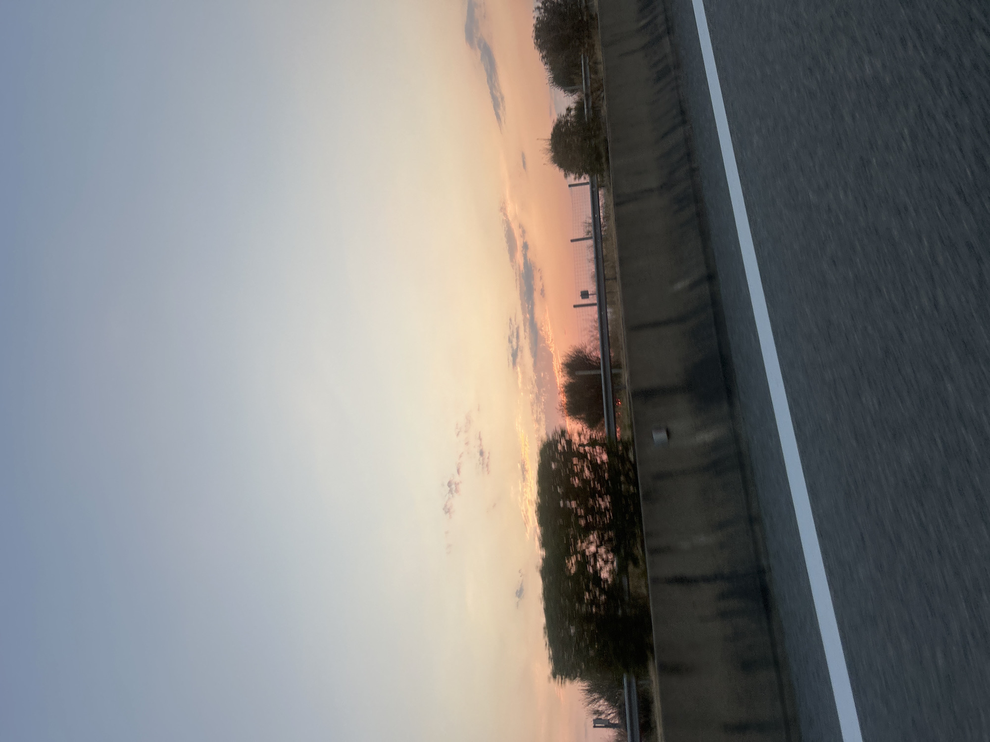 An image captures a view from a moving vehicle on a highway at dusk. The sky displays a gradient of soft blues and oranges, with sparse clouds tinged with pink. In the middle ground, a concrete barrier separates the highway from a landscape featuring low-lying vegetation, including bushes and some bare trees. A wire fence is visible behind the bushes. The highway pavement is dark gray with a prominent white line marking the lane. The overall scene depicts a roadside view during the golden hour. The location is the Municipal Unit of Etoliko, Greece.
