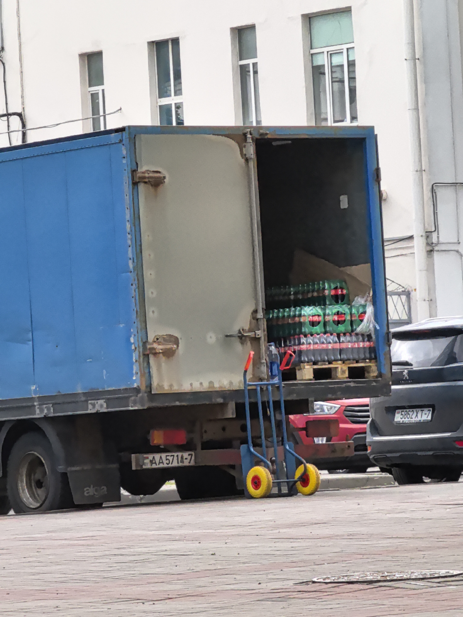 This media file shows the back of a blue delivery truck with its rear doors open, revealing stacks of soda bottles on pallets. The truck appears to be parked on a paved surface, possibly a street or a loading dock, with a building in the background. A red car is partially visible behind the truck, and a dark gray SUV is parked to the right of the truck, with a license plate that reads "5862 XT-7". A hand truck with yellow wheels is positioned in front of the truck's cargo. The lighting suggests it is daytime, and the weather appears to be overcast. No people are visible in the image. Text visible on the truck includes "alga" and a license plate that reads "AA 5714-7".
