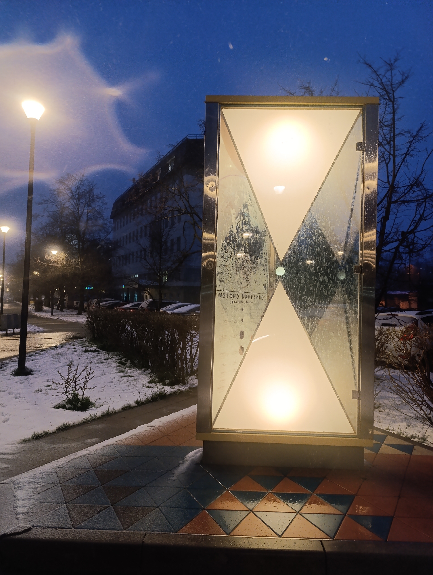 The image presents a quiet winter evening scene in Chernogolovka, Russia. In the foreground, to the right, stands a tall, modern, illuminated structure. It features polished metallic frames and frosted glass panels that create an hourglass shape with two brightly lit sections at the top and bottom. Its base is set on a distinctive mosaic of triangular tiles in terracotta and blue/grey, partially covered with a thin layer of snow.
To the left, a prominent streetlight emits a warm, bright glow, surrounded by noticeable light halos and flares, suggesting the presence of falling snow or ice crystals in the cold air. Further down the street, additional streetlights punctuate the scene. The ground, including a sidewalk and grassy areas, is lightly dusted with snow. Bare trees with intricate branches line the street and are silhouetted against the dark blue evening sky.
In the background, a multi-story building with numerous windows is visible, some reflecting faint light. Several cars are parked along the street, also bearing a light covering of snow. The overall time of day appears to be dusk or early night, characterized by the dark blue sky and active street lighting. On the reflective glass of the illuminated structure, mirrored Cyrillic text is visible, appearing as "МЕТСНО РАНРИЧОС", likely a reflection of a sign from an unseen source. The scene evokes a sense of cold tranquility.