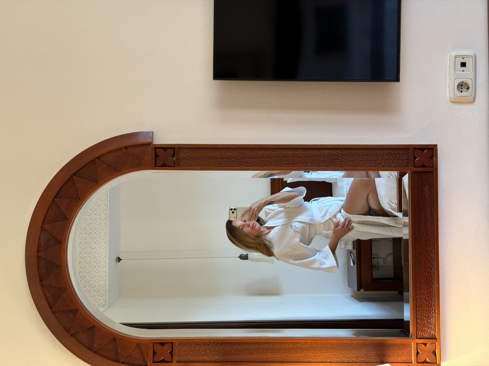The image depicts an indoor scene, likely a hotel room in Kuta, Indonesia. The primary subject is a woman, reflected in a large, ornately framed wooden mirror, taking a selfie with her smartphone. She is wearing a white terrycloth bathrobe, which clearly displays the text "GRAND" on it, indicating hotel branding. Her reflection shows her seated on a bed with white bedding, and a ceiling light fixture is also visible within the mirror's surface.
Above the mirror, a black flat-screen television is mounted on a light-colored wall. To the right of the television, an electrical outlet unit is visible, featuring a European-style power socket and what appears to be a data port. The mirror's frame is made of dark wood with intricate carved details, including repeating square motifs with an 'X'-like design in the corners and triangular patterns along its arched top. The scene suggests a well-appointed and comfortable accommodation setting. The lighting is characteristic of an indoor environment, with no discernible specific time of day or weather.