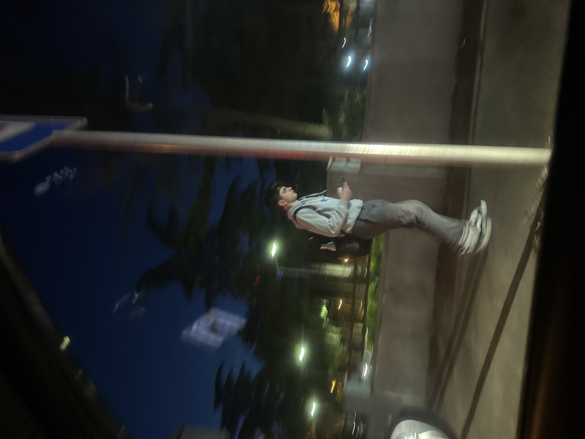 The image captures an urban street scene at night in Tbilisi, Georgia, likely viewed through a window, possibly from a moving vehicle, given the dark framing on the right and bottom, and blurred reflections. The central figure is a young male standing, dressed in a light grey zippered hoodie, dark grey pants, and white sneakers. He is holding a smartphone in both hands and appears to be looking at or interacting with the device, with his head tilted slightly upwards.
A prominent vertical metal pole with a reddish-brown band on it runs across the foreground/midground. To the left, a partial blue street sign is visible, but no specific text can be read. The background is dark, indicative of nighttime, featuring numerous elongated reflections of artificial lights (streetlights, building lights) and blurred silhouettes of trees, suggesting wet surfaces or strong reflections. The lighting is dim, relying on these artificial sources, creating a typical night-time urban ambiance. The young man seems focused and momentarily paused in his activity on the street.