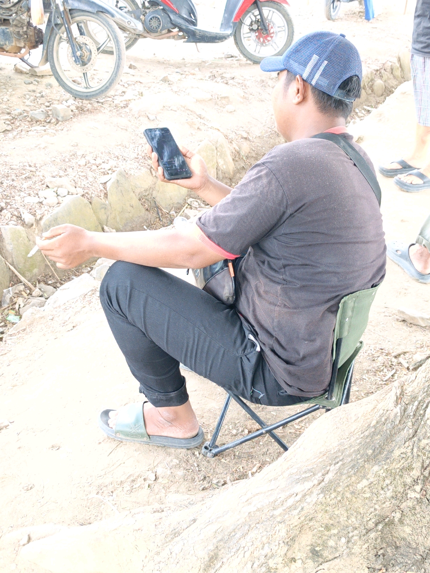 A man, wearing a dark t-shirt, black pants, and a blue baseball cap, sits on a folding green chair, holding a mobile phone and a cigarette in his right hand. He is outdoors, with a motorcycle visible in the background. The ground is a mix of dirt and rocks. The man appears to be in Gedung Nyapah, Indonesia.