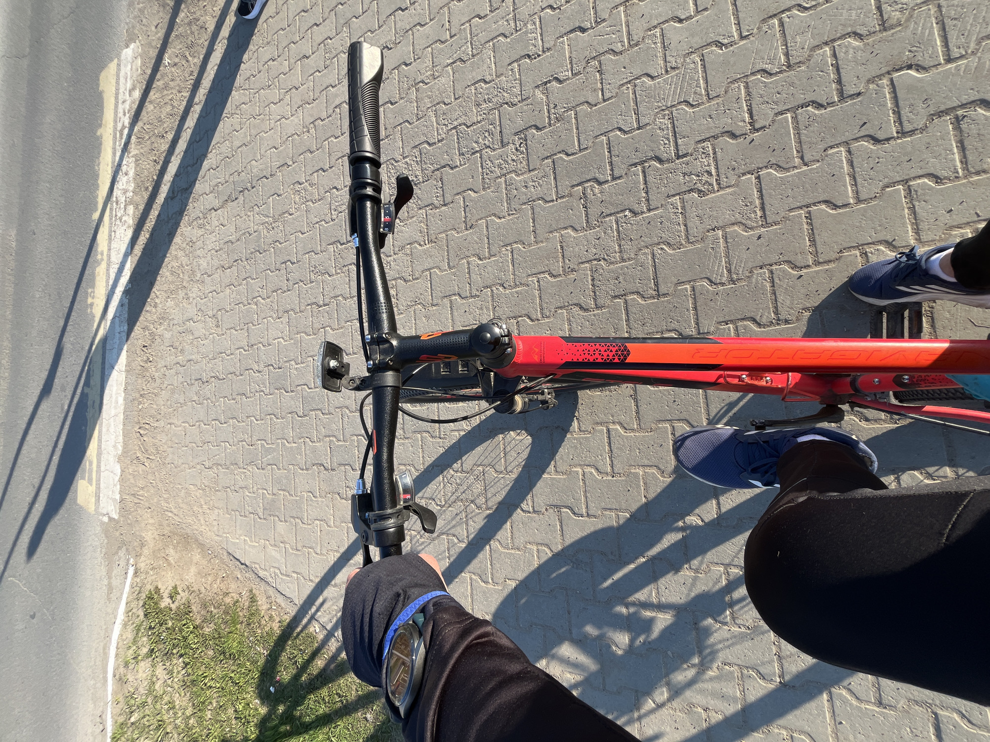 An eye-level, outdoor shot shows a portion of a red bicycle against a backdrop of brick pavement. The handlebars are prominent in the foreground. A person is visible with a gloved hand on one handlebar, wearing a watch. A leg and foot clad in blue athletic shoes and black pants are visible, with the feet on either side of the bike's frame. Part of the pavement is bordered by asphalt with a painted white line. Some sparse greenery is at the edge of the pavement. The location is the city of Chaikovsky, Russia.