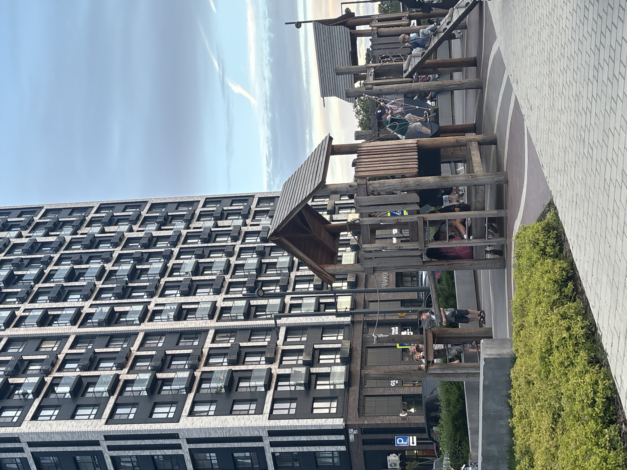 A vertical, eye-level shot captures a modern playground structure adjacent to a tall, dark-paned apartment building. The playground, constructed from rustic wooden beams and planks, features multiple levels, slides, and rope elements. Several people, including children and adults, are visible on and around the structure, seemingly engaged in recreational activities. The building's facade is characterized by a grid of dark windows, creating a repetitive and geometric pattern against the sky.
The time of day appears to be late afternoon or early evening, with a soft, diffused light casting subtle shadows. The sky is a pale blue with wispy white clouds and a hint of sunset hues. The overall atmosphere is calm and leisurely, suggesting a typical day in a residential area with amenities.
Notable details include the textured gray paving stones that form a pathway next to the playground and a dense green hedge bordering the scene. There are also glimpses of cars and street signs in the background, indicating an urban or suburban setting. Some text is visible on the building's lower floors, including what appears to be Russian signage like "ЧАЙКОФ" and "GU" and a parking symbol.
