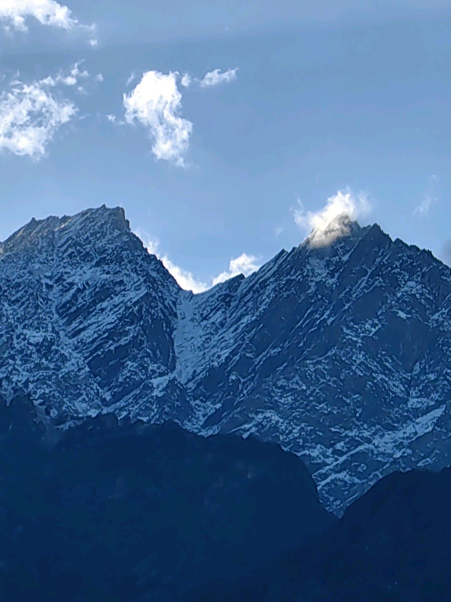 A wide shot captures a mountainous landscape under a bright blue sky dotted with wispy white clouds. Two prominent peaks dominate the center of the frame, their rugged surfaces covered in patches of snow, creating a stark contrast against the dark rock. A small cloud formation is visible near the summit of the right peak, partially obscuring it and illuminated by the sun. The lower portions of the mountains fade into a deep blue shadow, suggesting a vast, dark expanse below. The scene appears to be set in or near Vashisht, India, a region known for its Himalayan backdrop.