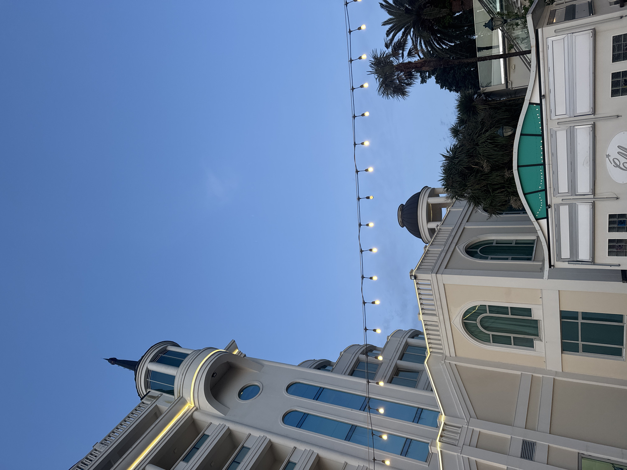An upward-facing, low-angle shot captures a serene late afternoon or early evening scene under a clear blue sky. The main subjects are two architecturally distinct buildings, connected visually by a string of illuminated warm-toned lights, and bordered by palm trees.
On the left side of the frame, a light-colored building rises, featuring a striking spire-like top, rounded windows, and pronounced horizontal decorative elements with subtle recessed lighting. The building's facade is smooth, possibly cream or light grey.
A horizontal strand of glowing string lights stretches across the upper mid-ground, providing a festive or inviting ambiance as twilight approaches.
To the right, another elegant, light-colored building is visible, characterized by arched windows and a dark-domed cupola or roof feature. Attached to this structure is what appears to be a commercial establishment, featuring a distinctive green, curved awning-like element and several rectangular panels. Partial white, stylized text that reads "ell" along with a small star or ornament, is visible on this section.
Several dark green palm trees are silhouetted against the clear sky in the upper right portion of the image, suggesting a warm climate. The scene is calm, with no human activity, focusing instead on the architectural details and the soft glow of the early evening lights.