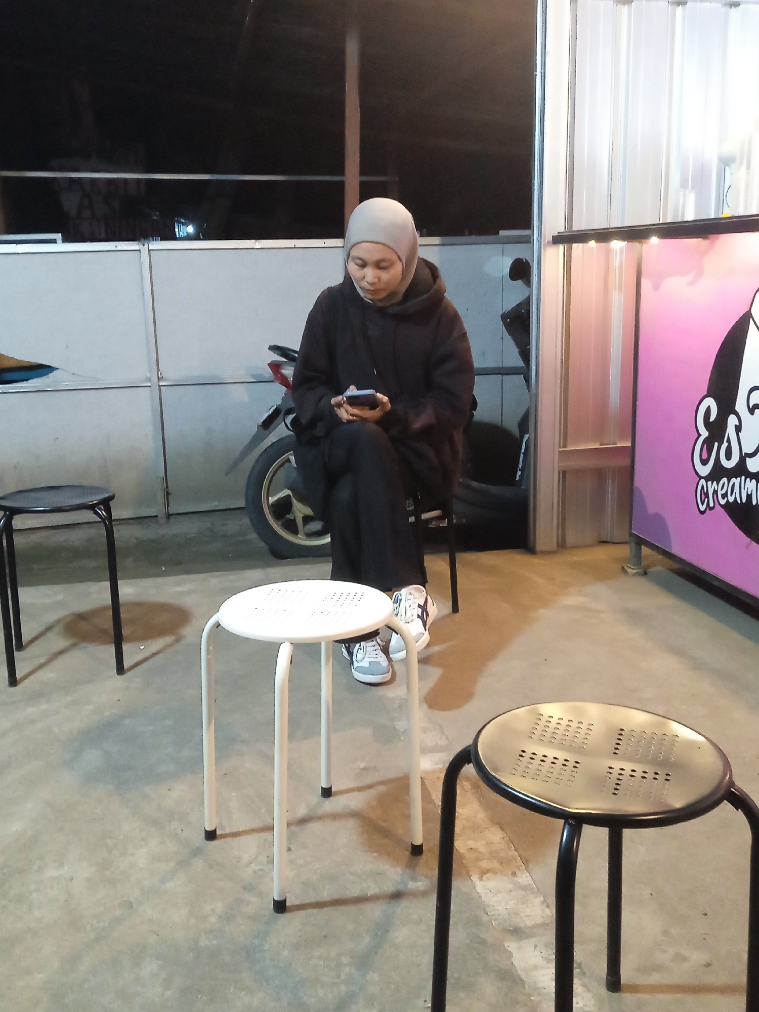 The image captures a casual night scene in Sekopek Wetan, Indonesia, likely an outdoor or semi-outdoor waiting area. The main subject is a person, appearing to be a woman, seated on a black stool. She is wearing a light grey hijab, a dark hoodie, and dark pants, with white sneakers that have grey and black details. Her attention is solely on her smartphone, which she holds in both hands.
In the foreground, there are two more stools: a white one closer to the viewer on the right, and another black one on the far left. The ground is a concrete surface. In the background, to the left of the woman, a motorcycle or scooter is parked. The wider background consists of dark, likely metallic or corrugated walls, suggesting a simple, possibly makeshift structure or a utility area.
To the right of the frame, there's a pink counter or booth with stylized text. Partially visible is "ES & CREAM," likely indicating an ice cream or creamy dessert stall. The lighting suggests it is nighttime, with artificial light illuminating the immediate area. The atmosphere appears calm, with the woman engrossed in her device.