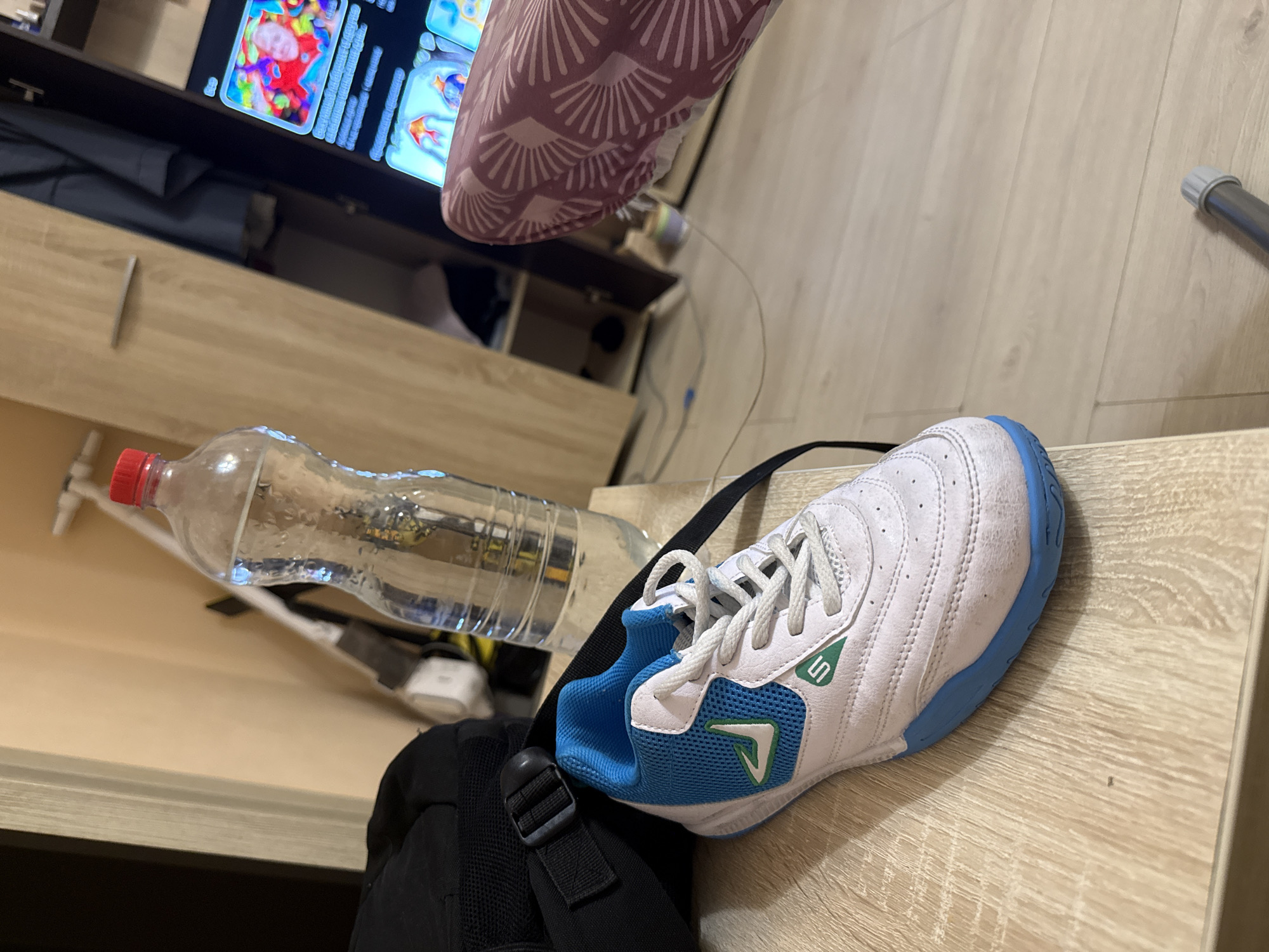 This image captures an indoor scene, likely a room or living area, in Tobolsk, Russia. The perspective is an angled overhead shot, focusing on items placed on a light-colored wooden surface, possibly a table or shelf.
In the foreground and midground, a white and blue athletic shoe is prominently displayed. It features white laces, a green stylized "S" or similar logo on its side, and a green V-shaped detail on the blue mesh portion. Next to the shoe is a clear plastic water bottle with a red cap, partially filled with water and showing condensation. A black backpack or bag, with a visible strap and buckle, is partially seen beneath the shoe and bottle.
The background reveals a television screen or monitor displaying colorful graphics, possibly from a video or game. One image shows what appears to be a multi-colored brain or abstract design, while another depicts goldfish. Unreadable text is visible on the screen, likely descriptions or titles. Below the TV, a wooden furniture unit, possibly a TV stand or cabinet, has an open drawer revealing dark fabric or items inside. A light-pink fabric item with a white fan-like or shell pattern is draped in the upper right, partially obscuring the TV. To the far right, a silver-colored pole or pipe is visible near the floor, and various wires indicate electronic connections in the background.
No people are present, and no specific activity is taking place; the items appear to be casually placed. The lighting is typical indoor illumination, offering no specific cues about the time of day or weather.