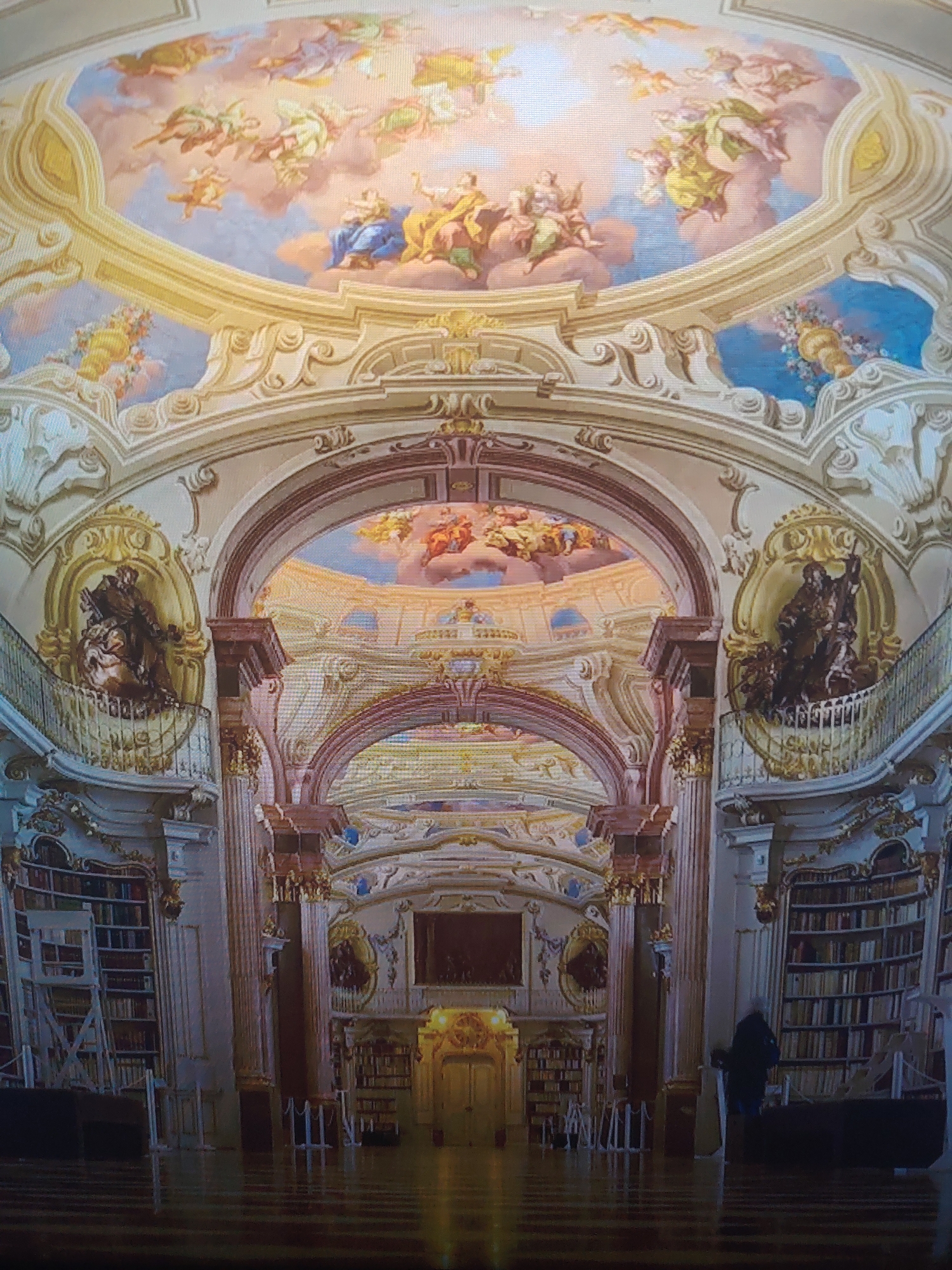 The image shows the interior of a large baroque library, possibly a monastery or abbey library. The location appears to be Unknown, Unknown. The architecture features ornate details, with frescoes on the ceilings depicting religious scenes. The walls are lined with bookshelves filled with books, with a few ladders visible to reach higher shelves. Two statues stand on balconies on either side. A single person stands towards the right side of the frame.