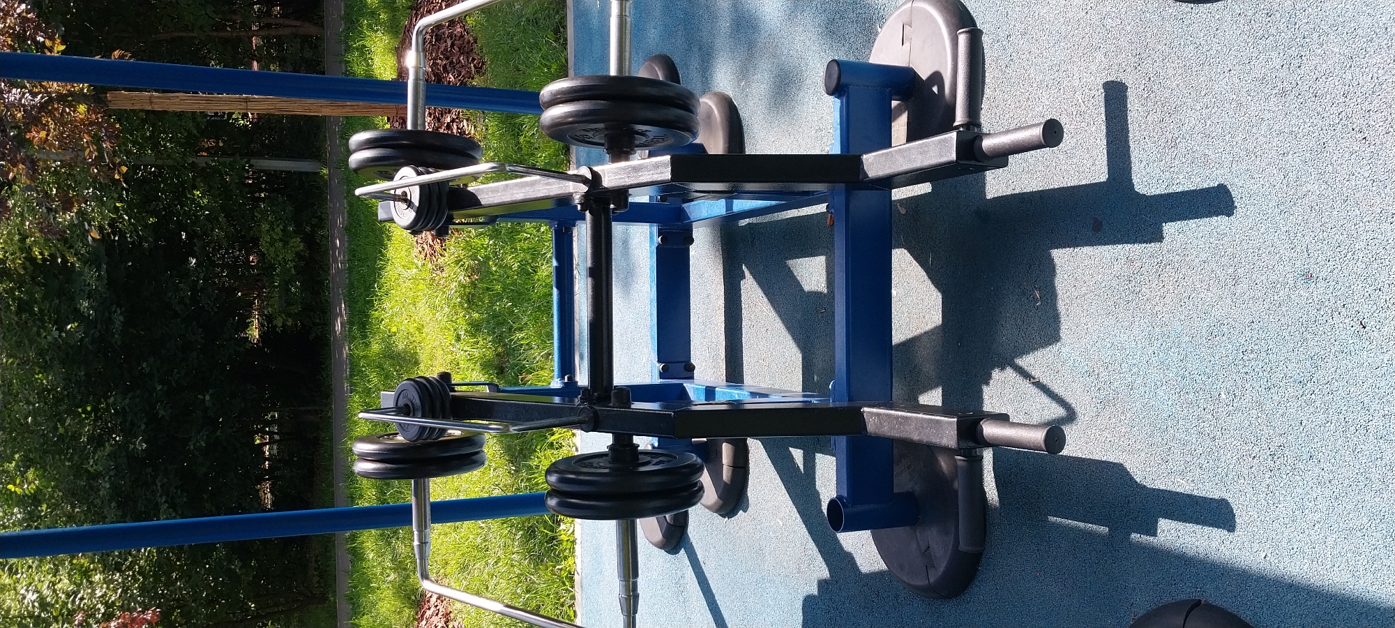 A weightlifting apparatus with barbells is situated on a blue, rubberized surface in an outdoor park setting. The apparatus is primarily blue and black, with silver metal components. Two barbells are loaded with black weight plates, stacked on each side of the central frame. In the background, green grass, trees, and bushes are visible under a bright sky. The image appears to have been taken on a sunny day, casting shadows from the equipment onto the ground. The location is identified as Moscow, Russia, suggesting this is an outdoor fitness area in a Russian park. There are no people visible in the image.