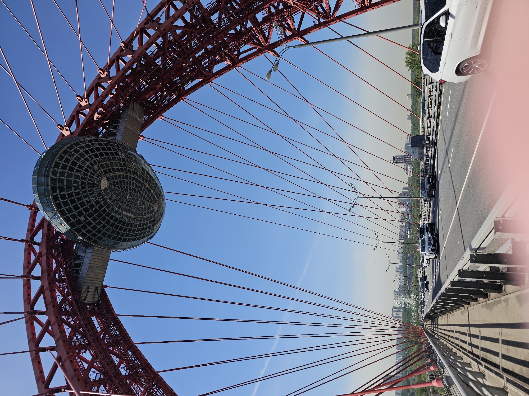 The image presents a low-angle view of the Zhivopisny Bridge in Khimki, Russia, with its distinctive red steel arch and the spherical viewing platform suspended at its apex. The platform is constructed of glass and metal gridwork. Red cable stays radiate outwards from the arch and the sphere. A concrete bridge deck runs parallel to the frame. A line of vehicles is visible on the bridge, moving along the road. The sky is blue, with some light haze near the horizon. Street lights are visible along the roadway. The city skyline is barely discernible in the background.