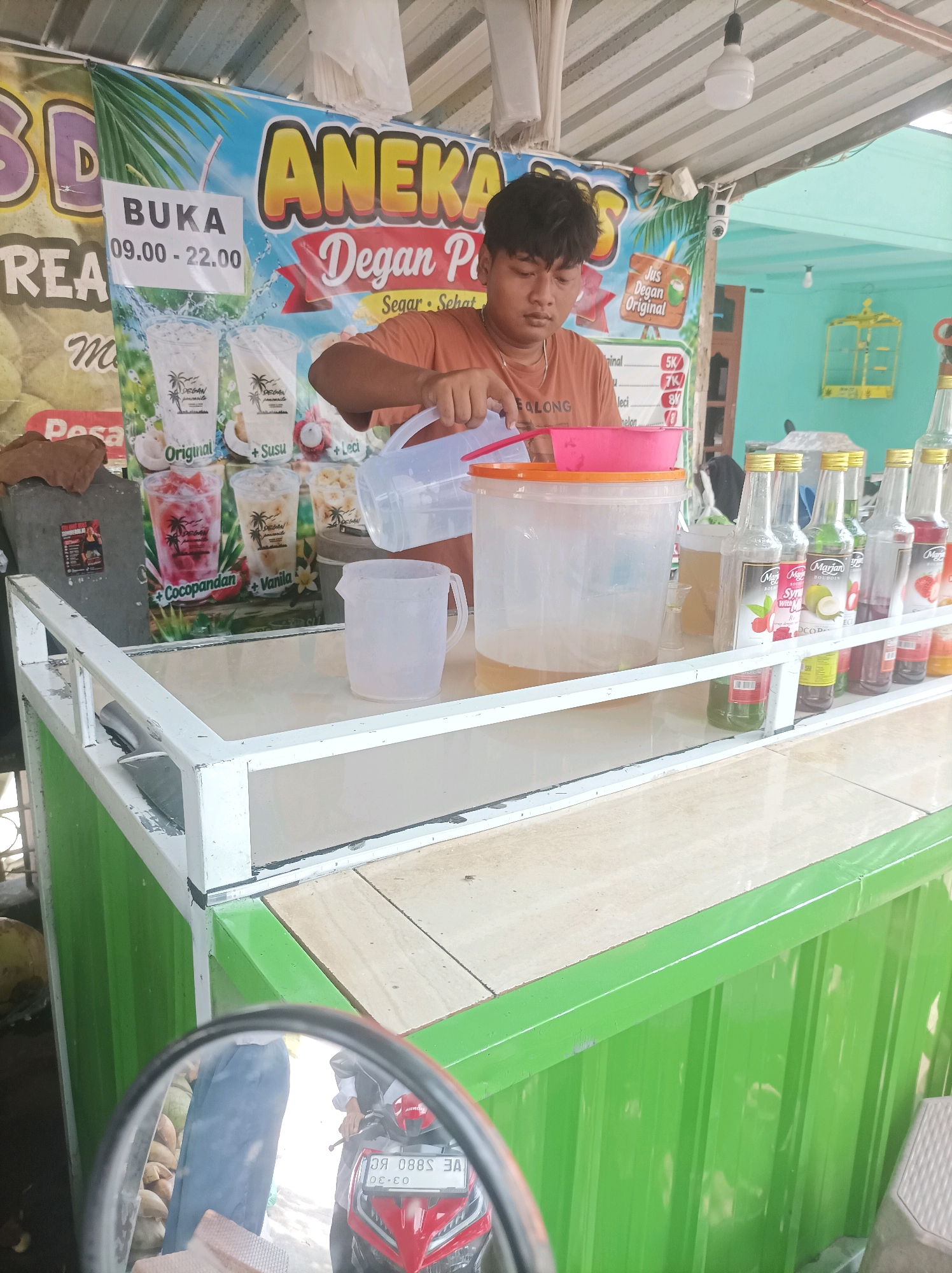A young male vendor with dark hair is preparing drinks at a roadside stall. He is wearing an orange t-shirt and is in the process of pouring liquid from a clear plastic container into a pink bowl. In front of him are several large plastic containers, one clear with some liquid and a pink lid, and another smaller, clear jug with a yellow liquid. Lined up on a shelf are numerous bottles of syrup, many with yellow caps and labels indicating flavors like strawberry and coconut. Behind the vendor is a large banner advertising "ANEKA Degan P" with images of drinks and text indicating opening hours of 09:00 - 22:00. The stall appears to be outdoors, with a corrugated metal roof and green corrugated metal siding visible. In the foreground, a motorcycle's side mirror reflects a person sitting on a motorcycle and some coconuts. The scene is likely located in Sumuragung, Indonesia, given the context.
