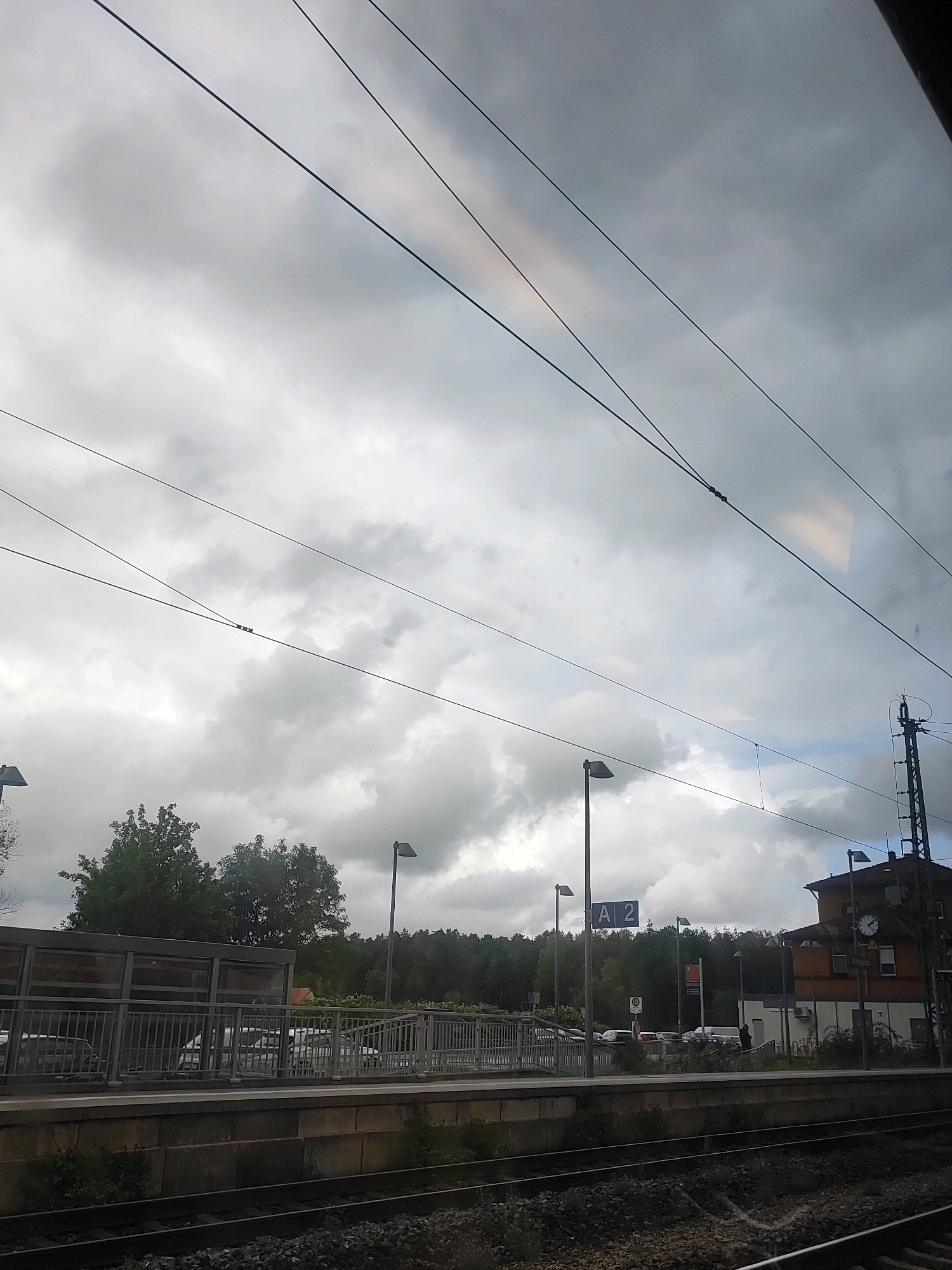 The image shows a train station platform in Petersaurach, Germany, on a cloudy day. Overhead power lines crisscross the sky, casting shadows on the scene. A train platform with a metal fence is visible, with cars parked behind it. A building with a clock is located at the far end of the platform. The sky is overcast with dramatic clouds. The overall atmosphere is somber and still, suggesting a quiet moment before or after a train's arrival or departure.