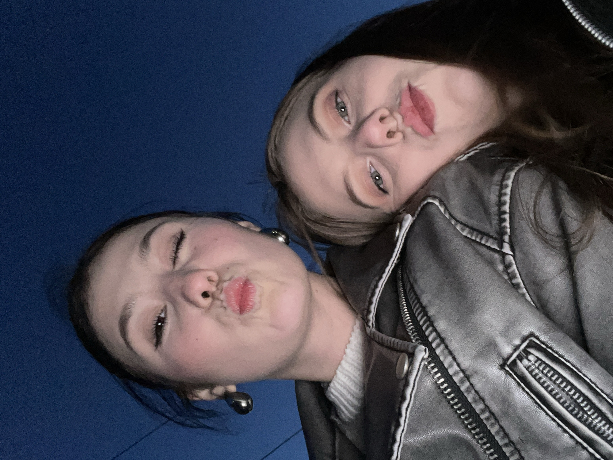 Two young women pose closely together. The woman on the left wears large, shiny, round earrings, and puckers her lips. The woman on the right is leaning her head onto the other woman's shoulder. She wears a silver-gray jacket and light makeup. A dark blue, possibly night sky, is visible in the background. The city of Фоки, Russia, is not discernible in the image.