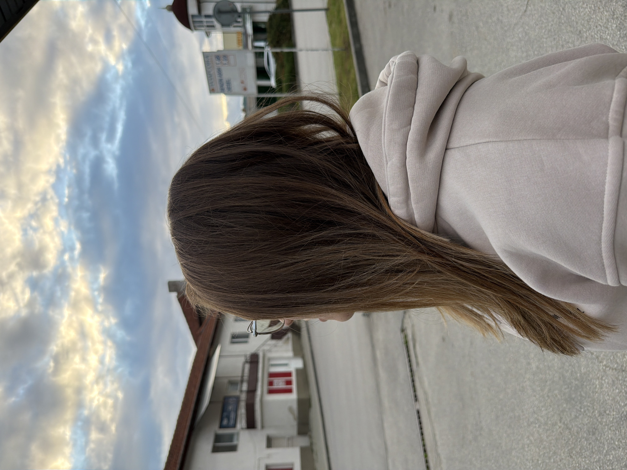 A woman stands outdoors on a paved street in Panagyurishte, Bulgaria, facing away from the camera. She wears a light beige hooded sweatshirt. Her brown hair is long and straight, falling down her back. She wears glasses. Behind her is a white building with a red roof and red and white window frames. A signpost is also visible. The sky is cloudy.