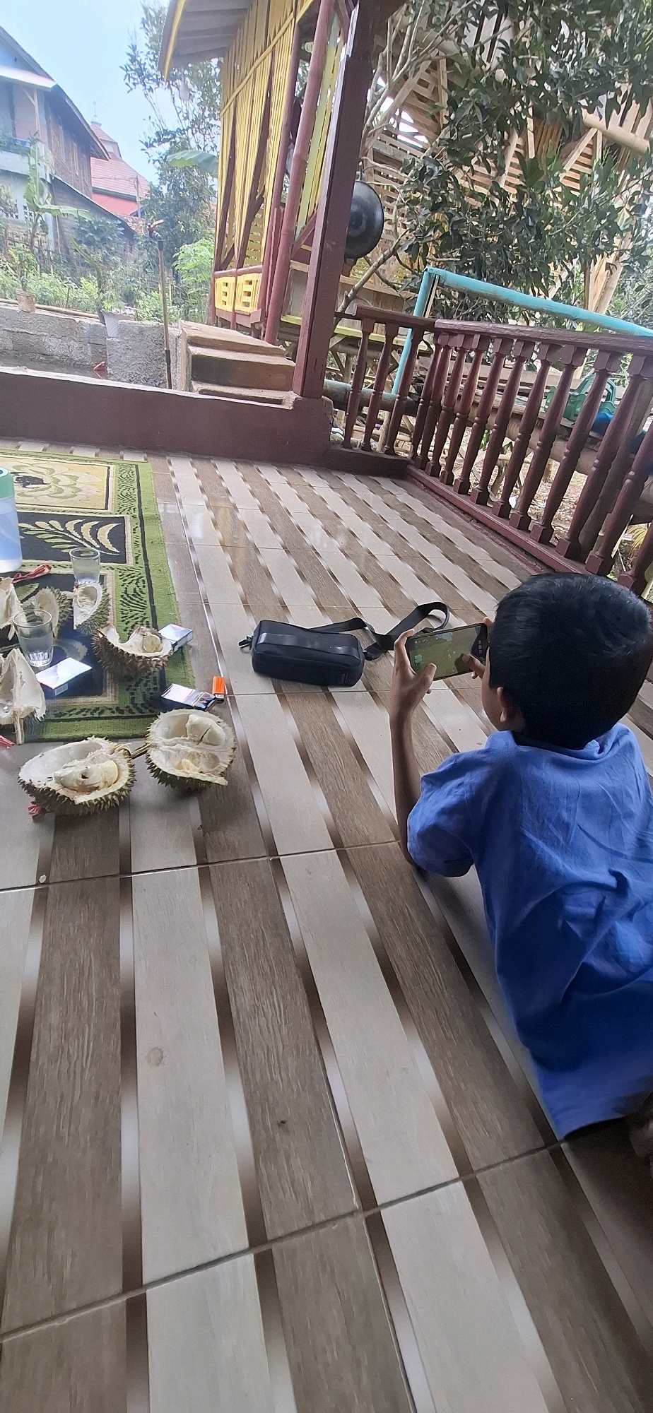 A young boy, wearing a blue shirt, lies on his stomach on a tiled porch in Cilaku, Indonesia. He is playing a video game on his mobile phone, which he holds horizontally. To his left, a black bag lies on the ground. The porch is decorated with a rug featuring a green and beige pattern, and scattered on the rug are several halves of durian fruit, some with visible flesh, along with glasses, cigarette packs, and a lighter. In the background, a wooden structure with yellow and red accents is visible, as well as trees and a glimpse of houses.