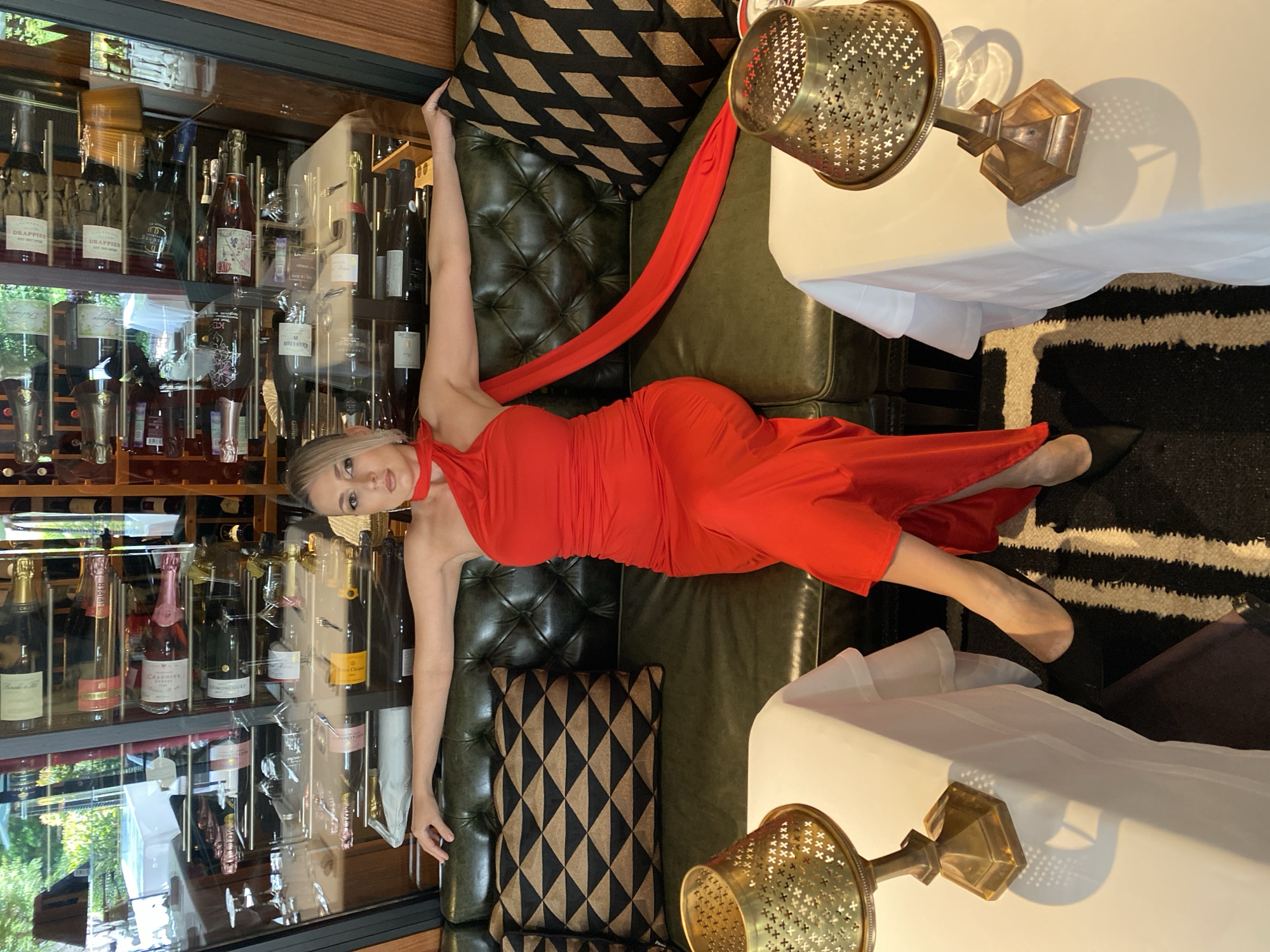A woman wearing a red dress is seated on a dark green tufted leather couch. She is positioned in the center of the frame with her arms outstretched, leaning back on the couch. Behind her is a large glass-fronted wine rack filled with numerous bottles of wine and champagne. The wine rack is illuminated, and the bottles are organized on multiple shelves. To her left and right are small, round tables covered with white tablecloths, each with a decorative golden lamp. The floor is covered with a black and white striped rug. The setting appears to be an upscale restaurant or bar. The woman's facial features include blonde hair pulled back, makeup, and she is looking slightly to her right. She is wearing black high heels.