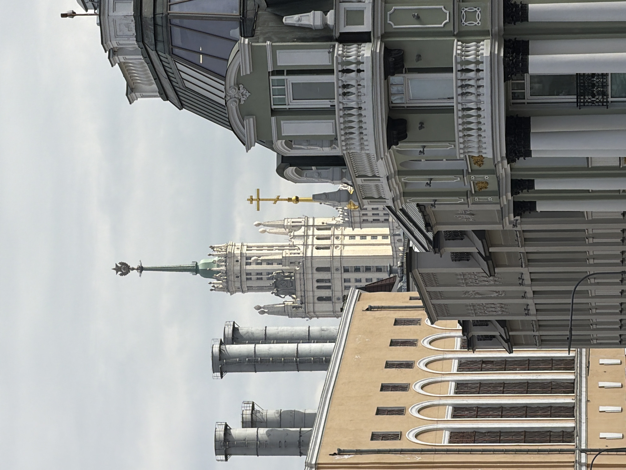 The image shows buildings in Moscow, Russia. In the center stands a Stalinist skyscraper, likely the Ministry of Foreign Affairs. In the foreground, there are industrial chimneys, and on the right, an older building with arched windows. Above, there is a building with elaborate baroque-style details. The sky is overcast.
