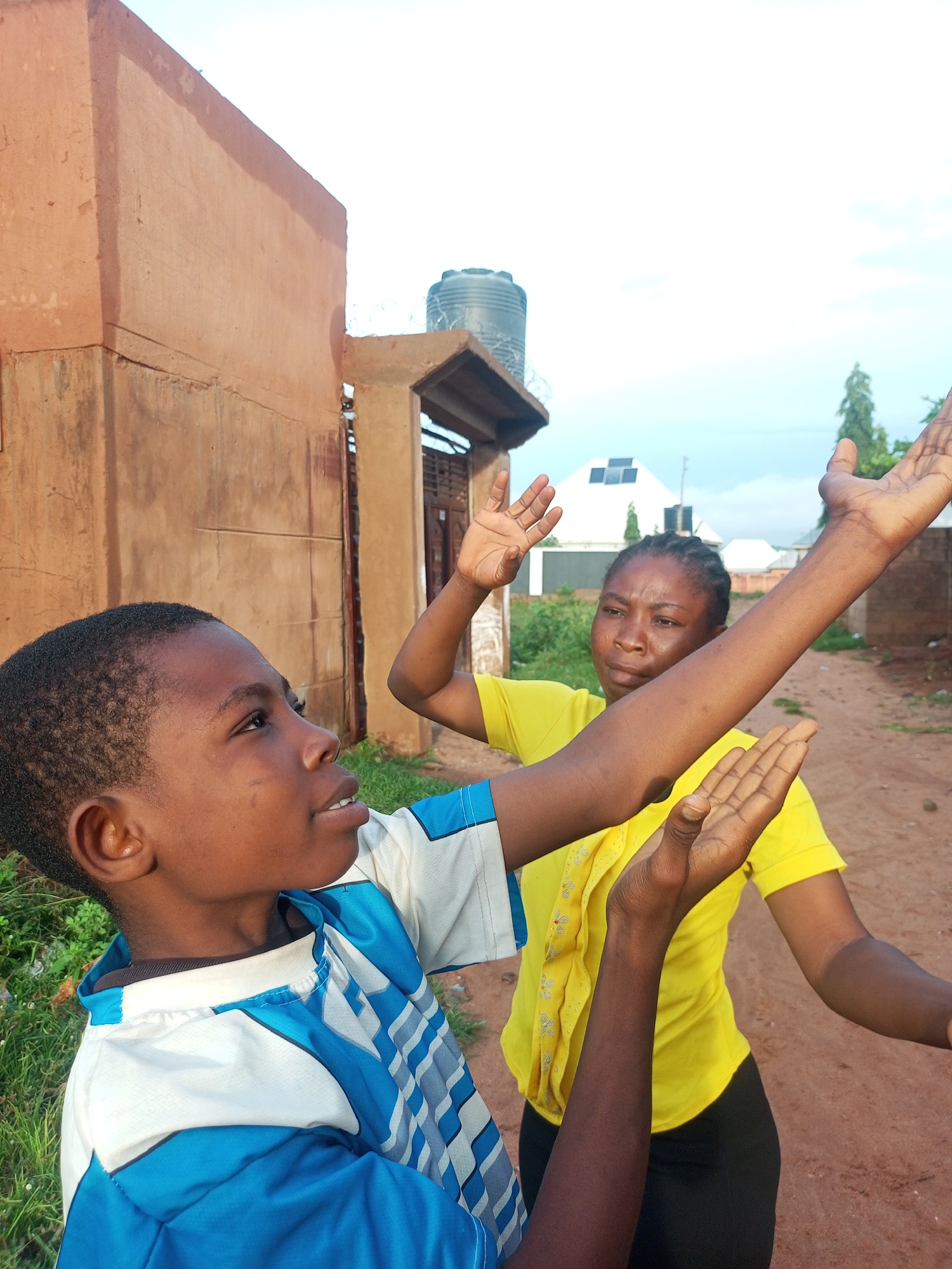 In Tempure, Nigeria, a young boy in a blue and white shirt is seen from the chest up, looking to his left with his mouth slightly open. He has his left arm raised, palm facing forward. Behind him, a woman wearing a yellow t-shirt and black pants is visible from the chest up. Her right arm is raised and extended, palm facing forward, while her left arm is bent at the elbow, with her hand near her face. She is looking downwards. They appear to be interacting in an outdoor environment with a dirt path and a terracotta-colored building to the left. A dark water tank is visible on top of a structure in the background, along with a white building with solar panels on its roof. The sky is bright and overcast.