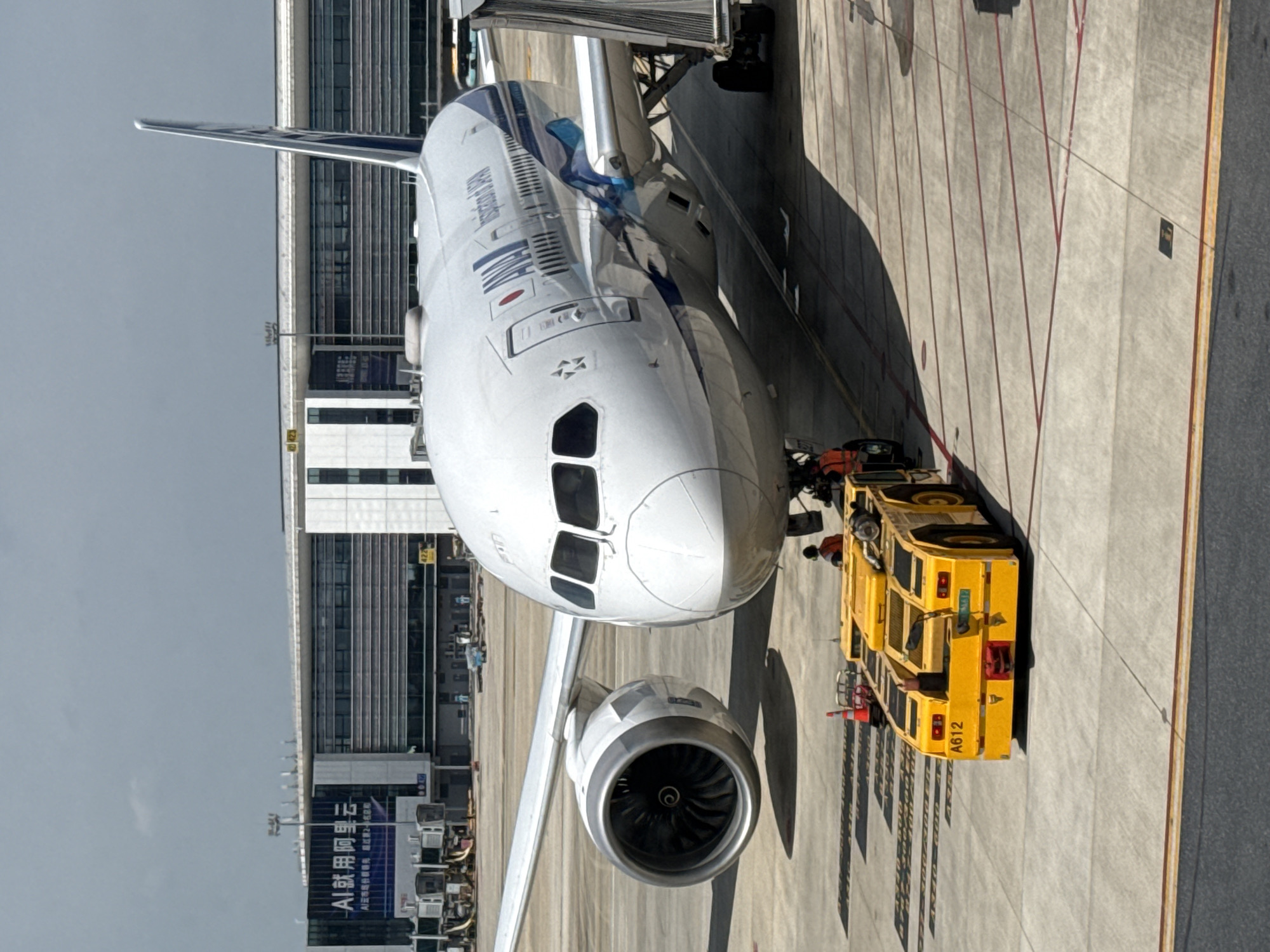 An ANA (All Nippon Airways) Boeing 787-9 Dreamliner is parked at an airport gate. The aircraft is white with blue and light blue accents. The registration number is not clearly visible. A yellow baggage tug, identified by the markings "A612" and "HNAH," is positioned in front of the aircraft's nose gear. Two individuals in orange vests are visible near the tug. In the background, the terminal building of Xiaoshan District International Airport in Hangzhou, China, is visible. The building features large glass windows and signage in Chinese. The sky is overcast. The scene appears to be during daytime.