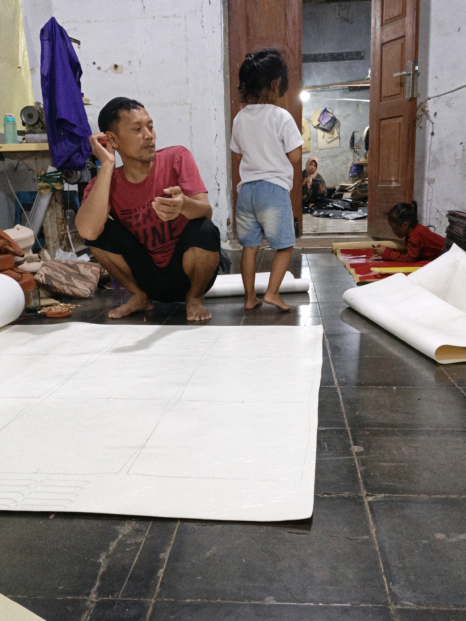 A man in a red shirt and black shorts is squatting on a tiled floor next to a large roll of white paper. He is holding a small red object in his right hand and appears to be talking or listening. A young girl in a white t-shirt and denim shorts stands facing away from the camera, looking into an open doorway. Another young girl in a red shirt is lying on the floor in the background, near more rolls of paper. A woman in a headscarf is visible through the doorway, working at a table. The scene appears to be a workshop or home in a modestly furnished room with a concrete floor and white walls. The overall impression is of a domestic or small business setting. The location is the city of Mangli, Indonesia.