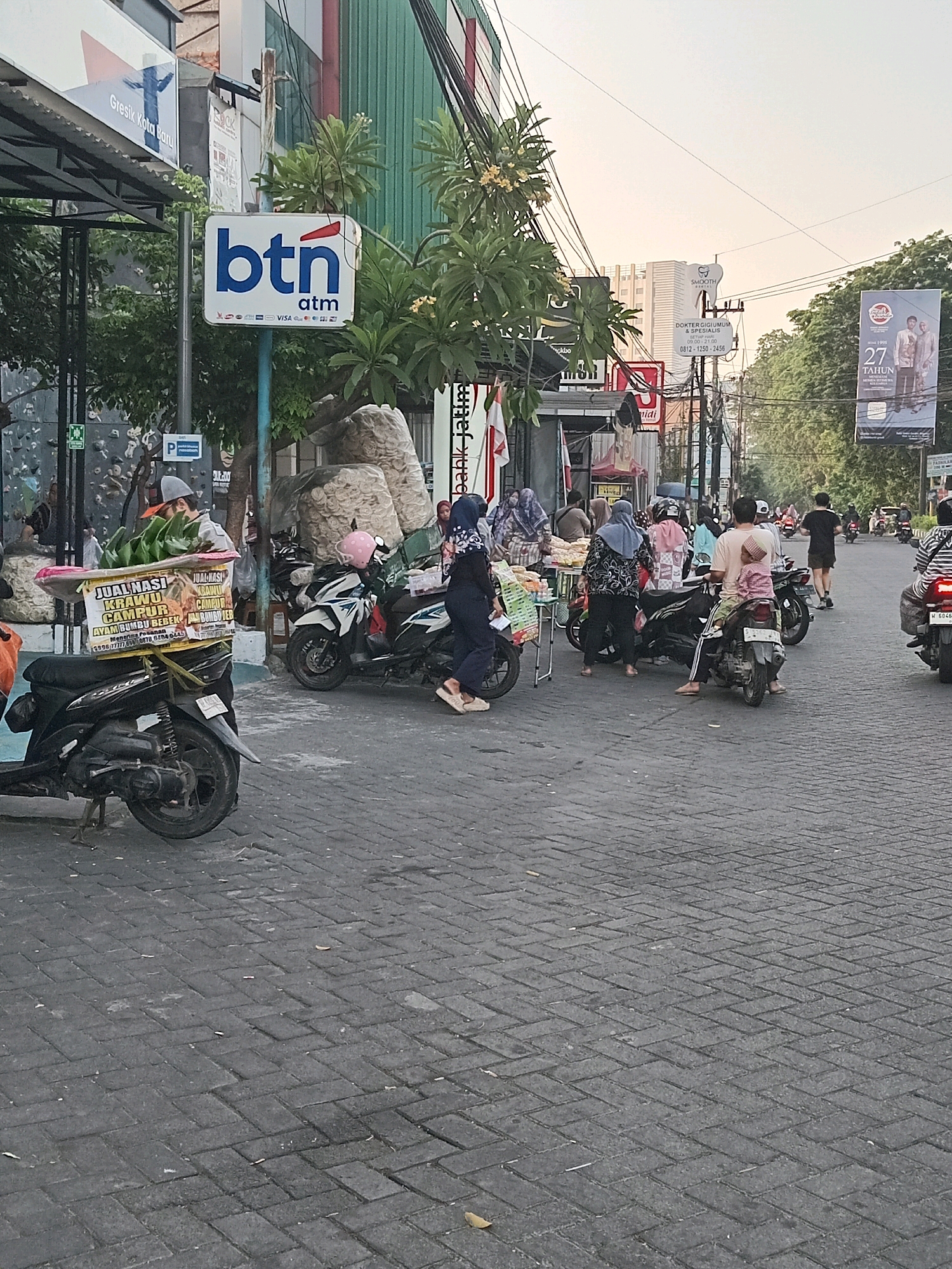 In Randuagung, Indonesia, the image captures a bustling street scene, likely during the day given the lighting. A paved street with interlocking bricks forms the ground. On the left, a stall is set up, displaying food items, and a man in a grey cap is seen attending to it. Several motorcycles are parked or being ridden through the area.
People are engaged in various activities, including shopping and commuting. Some individuals are dressed in traditional Indonesian attire, like hijabs. In the background, buildings and trees line the street, suggesting an urban or semi-urban environment. A prominent BTN (Bank Tabungan Negara) ATM sign is visible, indicating a financial institution's presence. Further down the street, a large banner advertises "27 TAHUN" (27 Years), possibly for a business or event. The overall atmosphere appears dynamic and representative of daily life in this Indonesian locale.