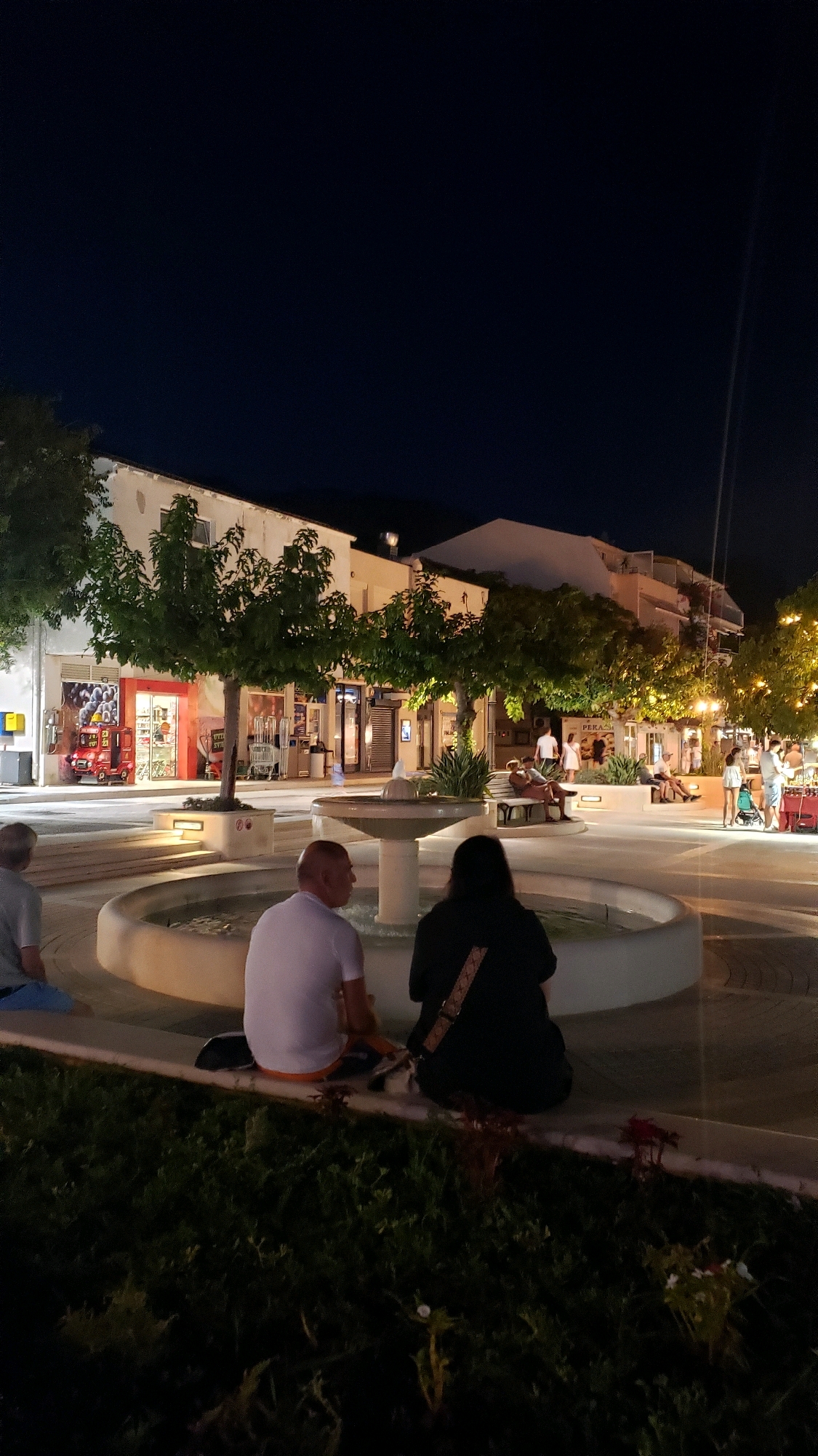 The image depicts a nighttime scene in a public square in Općina Tučepi, Croatia. A group of people are casually gathered around a fountain in the foreground. The square is illuminated by streetlights and lights from surrounding buildings, which appear to be shops or residential units. Trees line the perimeter of the square, and some have decorative lights strung between them. The architecture suggests a Mediterranean or coastal influence. The overall atmosphere is relaxed and social, with people enjoying the evening.