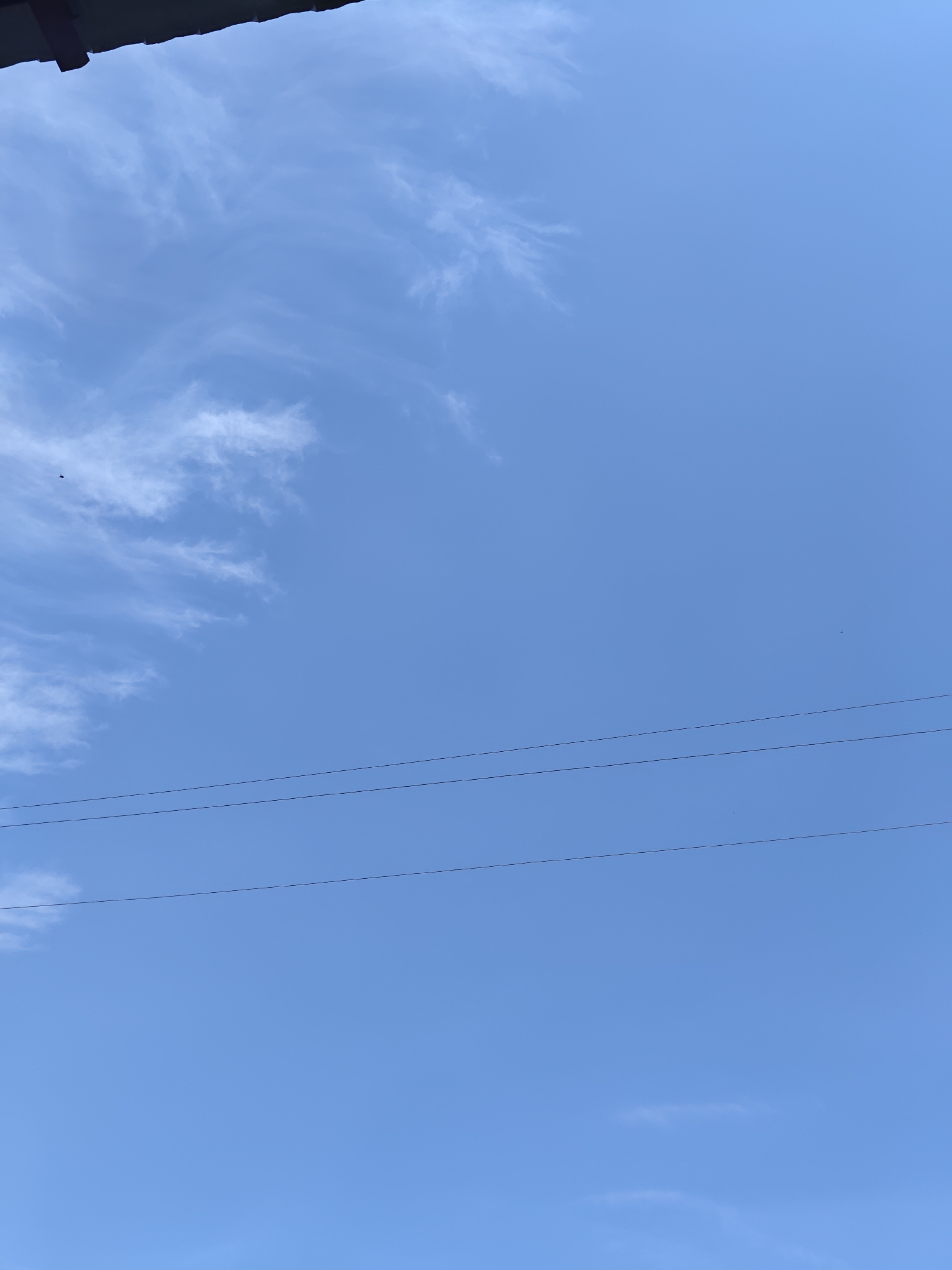 A clear blue sky with scattered wispy white clouds is visible. In the upper left corner, a dark, angled object, likely the edge of a roof or overhang, is present. Three parallel thin black lines, presumably power or telephone wires, stretch horizontally across the sky. The scene appears to be an outdoor view, possibly from a residential area in the city of Platoskino, Russia, given the context provided. There are no people or animals clearly discernible in the image.