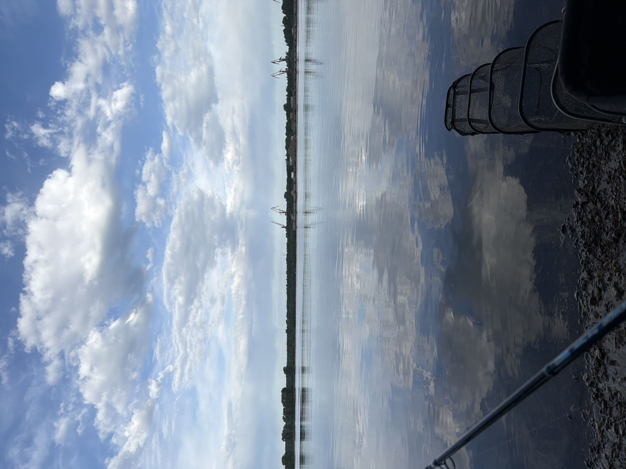 The image depicts a body of water, likely a river or large lake, reflecting a cloudy sky. The location is Chaikovsky, Russia, evident from the presence of industrial cranes along the shoreline. A fishing net is visible at the edge of the water, suggesting someone is fishing. The foreground shows a rocky shoreline and part of a fishing rod. The overall environment appears calm and scenic, despite the industrial elements in the background.