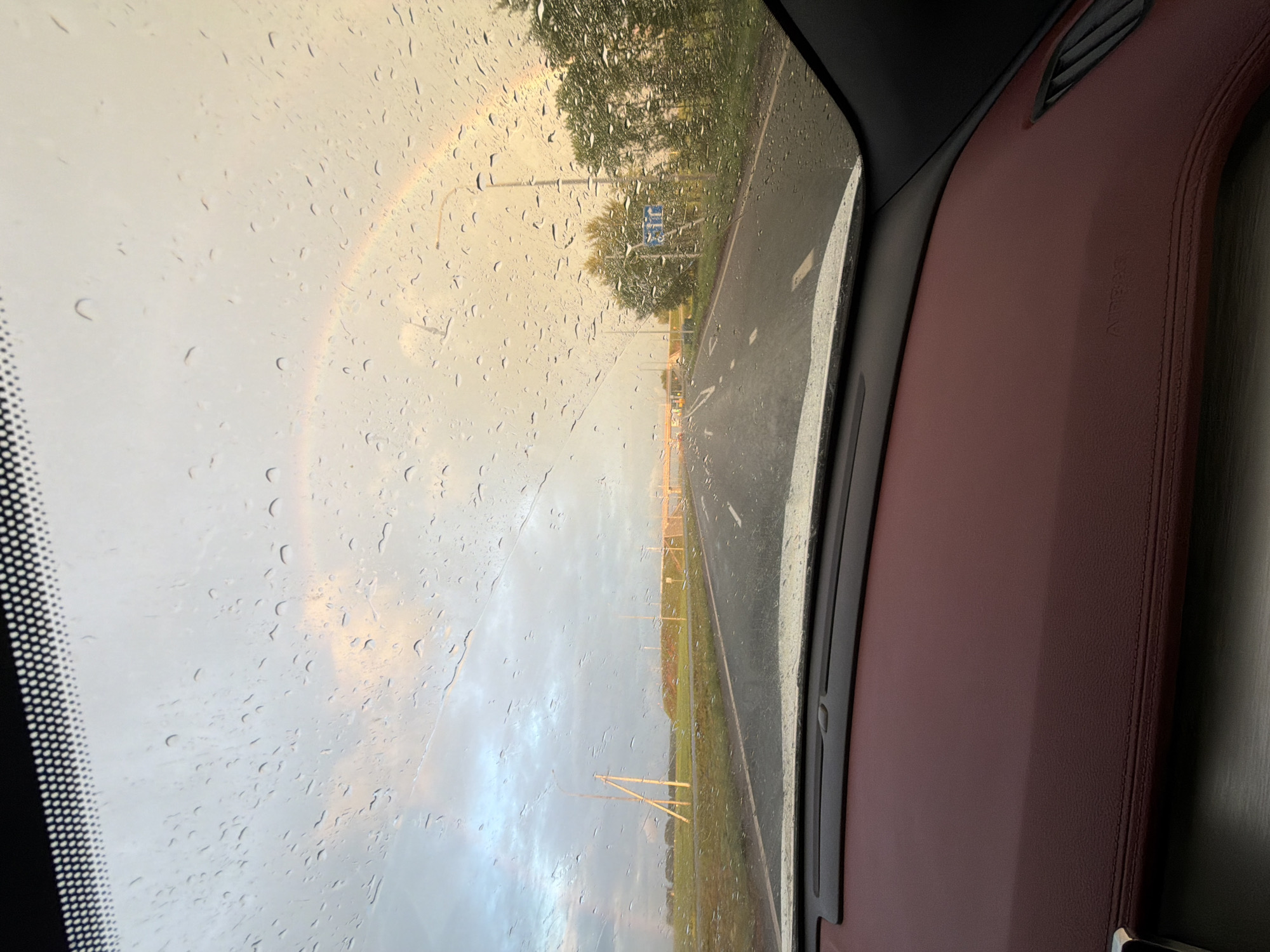The image captures a view through a car windshield, displaying raindrops and a partial rainbow in a sky with soft, warm lighting. The road ahead appears wet, with white lane markings visible. On the sides of the road, there are fields, trees, and utility poles, along with a road sign indicating public transport facilities. The interior of the car shows a dark dashboard with burgundy leather accents. The scene suggests a drive through a rural or suburban area during or shortly after rainfall, under a dramatic sky. The location in Таймурзино, Russia, is mentioned, providing context to the geographical setting.