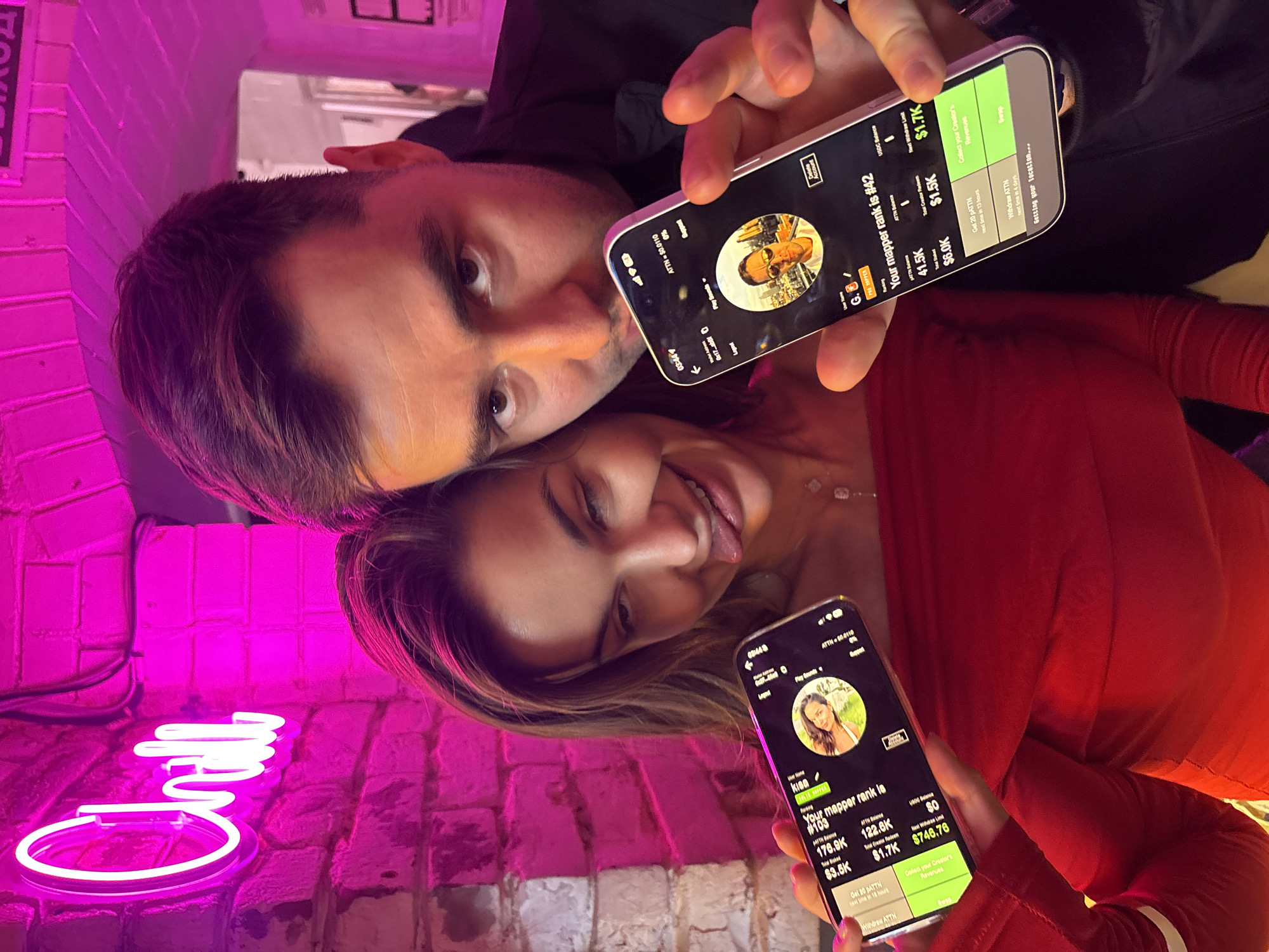 A selfie depicts a man and a woman in an indoor setting, potentially Krasnoyarsk, Russia. The man, fair-skinned with short dark hair, has one eye visible, looking into the camera while holding his phone. The woman, likely fair-skinned with light brown hair, sticks out her tongue. She is holding her phone. Each phone shows a different screen of an app, and they are overlapping each other. The woman wears a red dress, while the man wears a dark jacket. A pink neon sign reading "Chill" is visible.