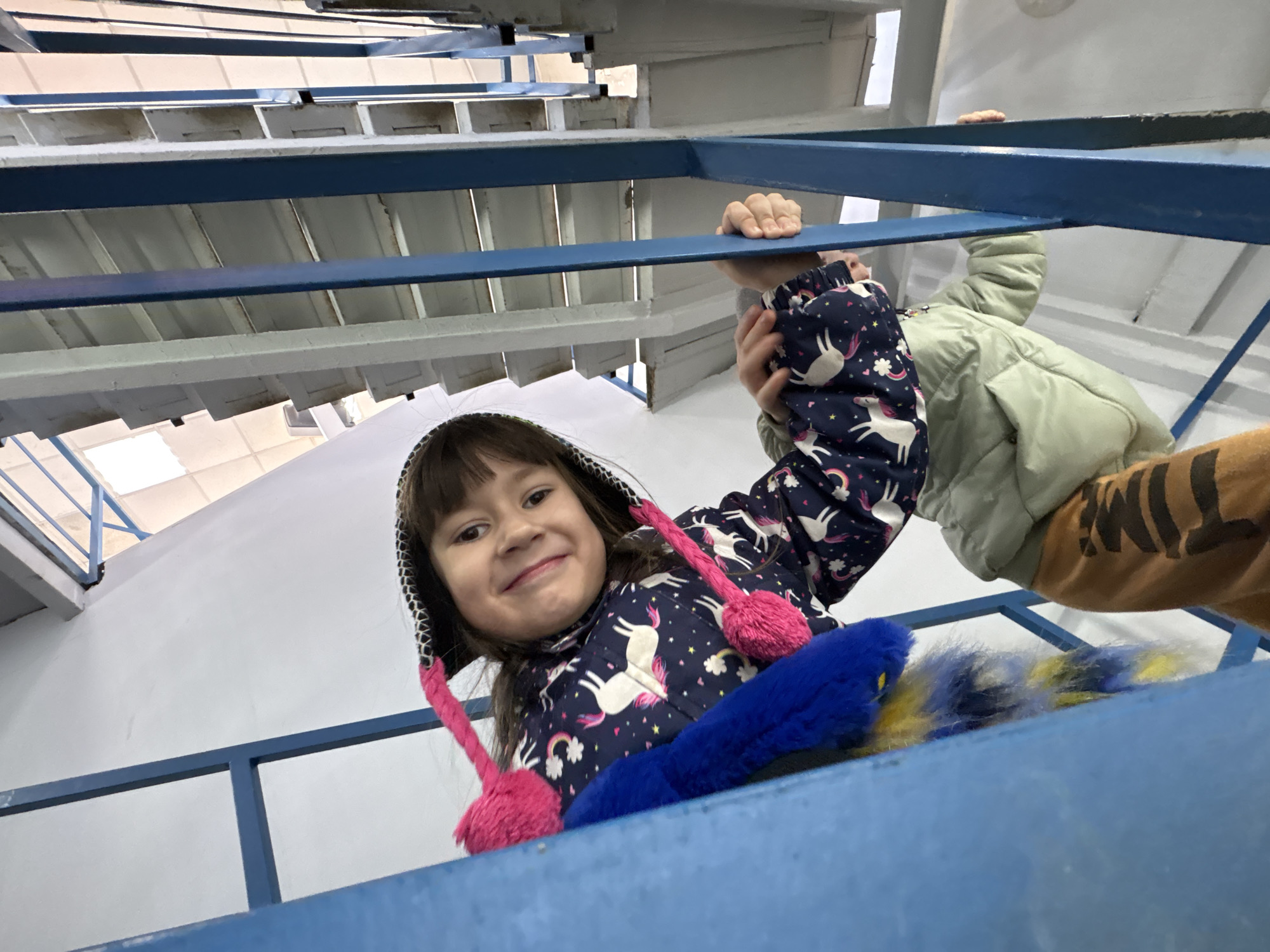 The scene depicts two children on an indoor staircase with blue metal railings, captured from a low-angle perspective looking upwards. The environment features white walls and light-colored stair treads visible in the background. In the foreground, a young child with dark hair and bangs smiles directly at the camera. This child wears a dark blue jacket adorned with white unicorn, rainbow, and star patterns, and a hood featuring a black-and-white checkered lining and two pink pom-poms attached via cords. A blue furry object with yellow accents is partially visible to the left of the child. Below and slightly behind this child, a second child, wearing a light green puffy jacket and brown pants with black horizontal stripes, grips the blue metal railing with both hands. Both children are positioned as if ascending the staircase. No specific identifying markers for a location within Izhevsk, Russia, are present in the image.