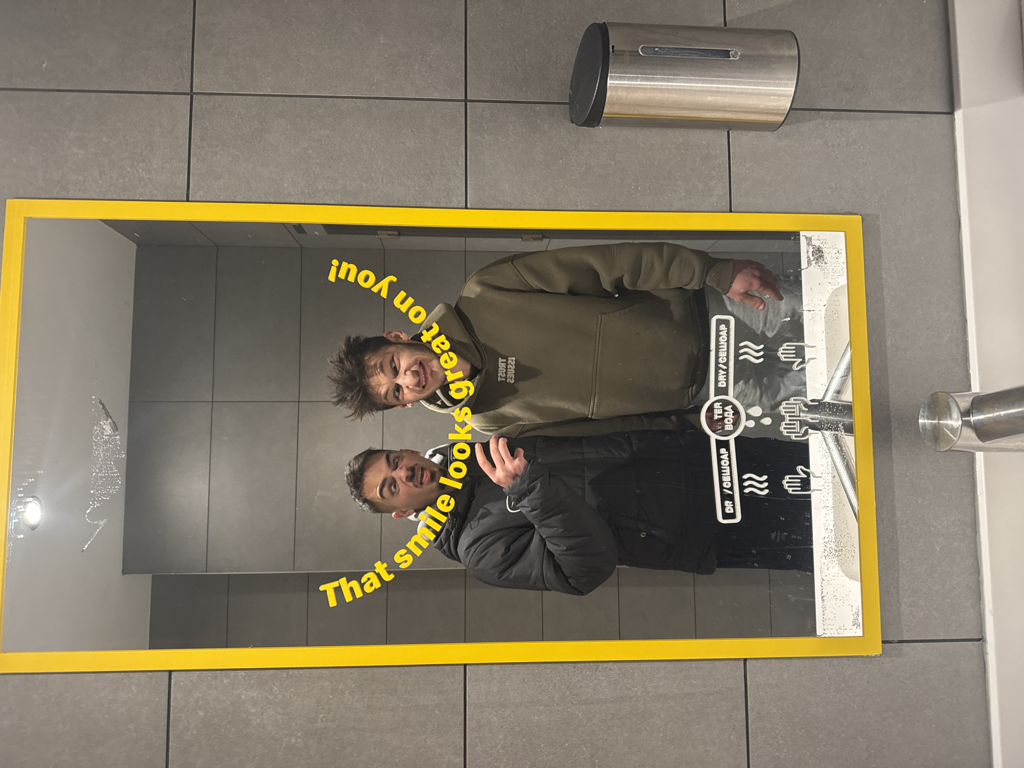 The image shows a mirror with a yellow frame in a public restroom. Two young men are reflected in the mirror, one wearing a black jacket and the other a khaki hoodie. Text superimposed on the mirror reads, "That smile looks great on you!" The mirror also contains instructions for drying or washing hands, with symbols for water and air. Above the mirror is a stainless steel soap dispenser. The wall behind the mirror is tiled with gray square tiles. The image was likely taken in Sofia, Bulgaria, based on the Cyrillic characters in the "DRY/СЕШОАР" instructions.