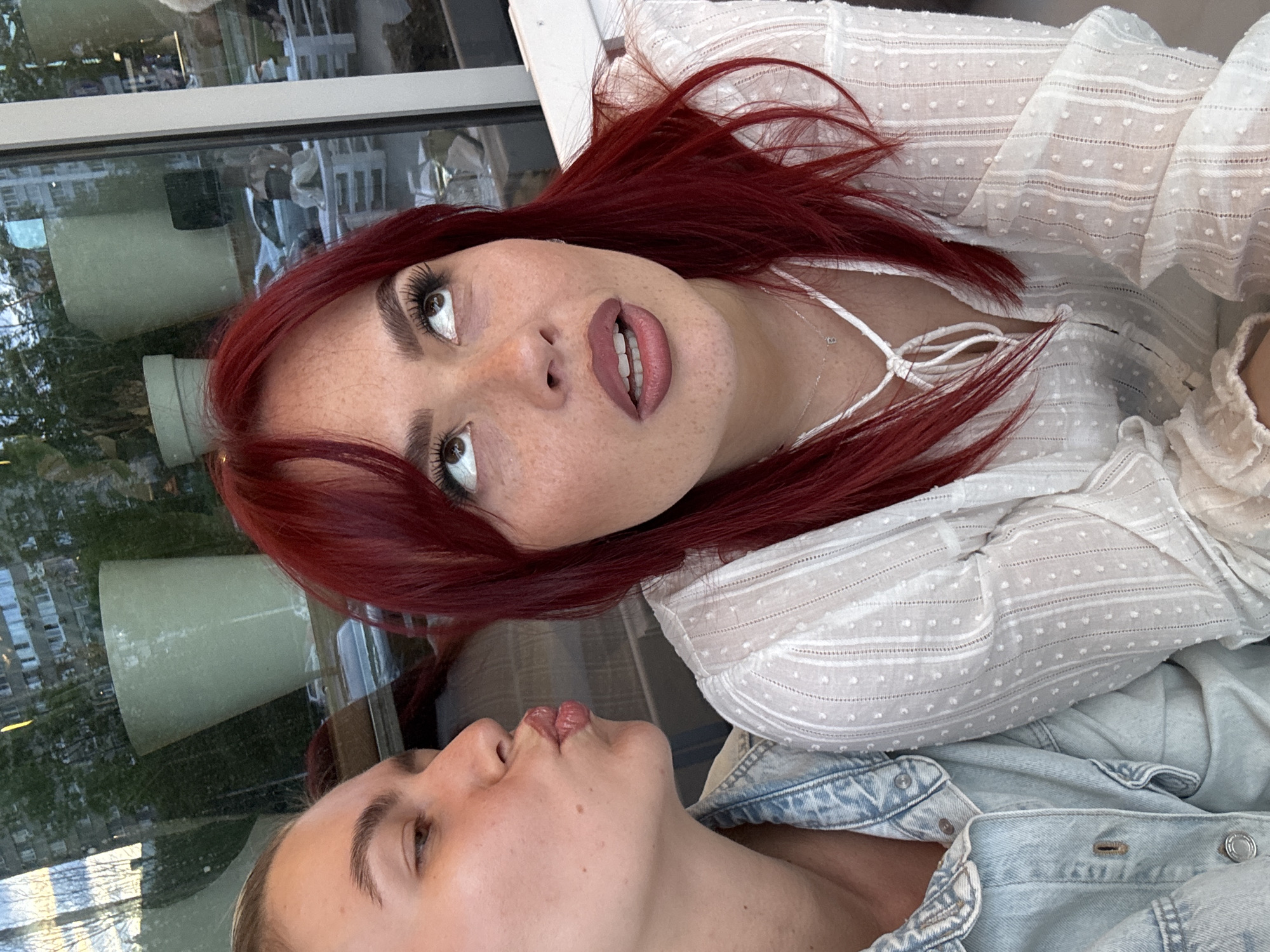 Two women are depicted in a close-up, outdoor, daytime shot. The woman on the left has blonde hair pulled back and is wearing a denim jacket. She is puckering her lips, as if to give a kiss. The woman on the right has bright red hair and is wearing a white textured blouse with a tie detail at the neck. She is looking upwards and slightly to her right, with her mouth open. Her expression appears surprised or thoughtful. Both women have makeup on, with the woman on the right sporting prominent eyelashes and freckles. The background is blurred and appears to be a reflection in a window, showing a cityscape with buildings and some greenery. The overall tone of the image is candid and personal, possibly taken during a social outing.