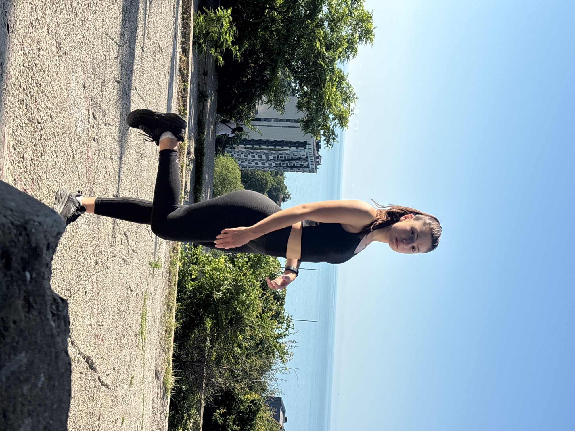 A young woman wearing a black sports bra and black leggings stands on a cracked concrete surface, one leg bent at the knee and lifted behind her. She has her hair in a ponytail and is looking towards the camera. The background shows a clear blue sky, a body of water, lush green trees, and a tall, modern building. In the distance, a person is seated on a stone ledge. The scene appears to be outdoors, possibly in a park or elevated viewpoint overlooking the sea, consistent with locations found in Varna, Bulgaria.