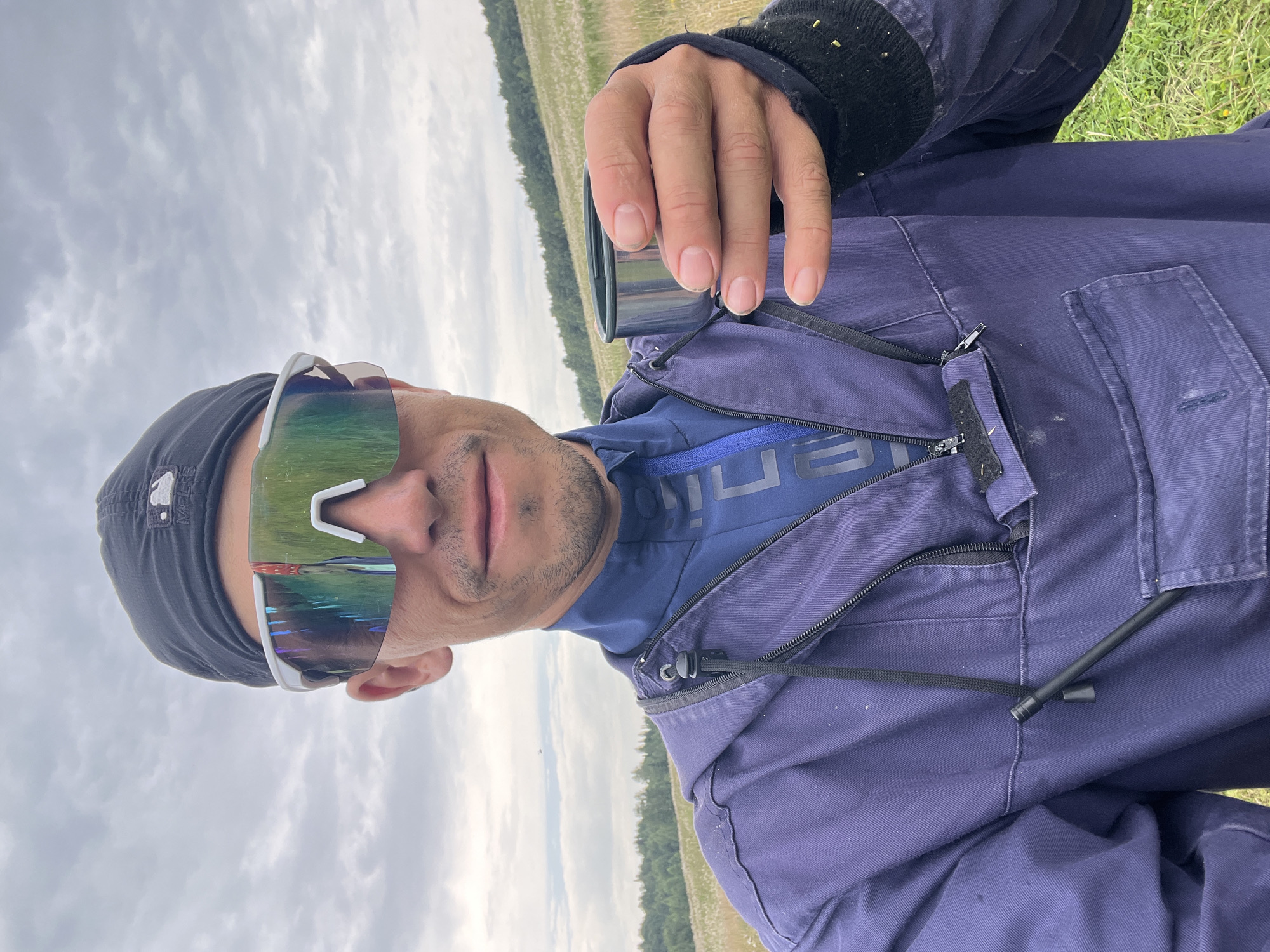 In an outdoor, rural setting resembling a meadow or field, a person is taking a selfie. They are wearing a black baseball cap and large, mirrored sunglasses with a white frame. The person is holding a small metal cup in their right hand and is wearing a dark blue hooded jacket over a blue athletic shirt. The background consists of a grassy field with scattered white flowers, a treeline in the distance, and a cloudy sky. There is no indication of the specific location within Auk-Bulyak, Russia.