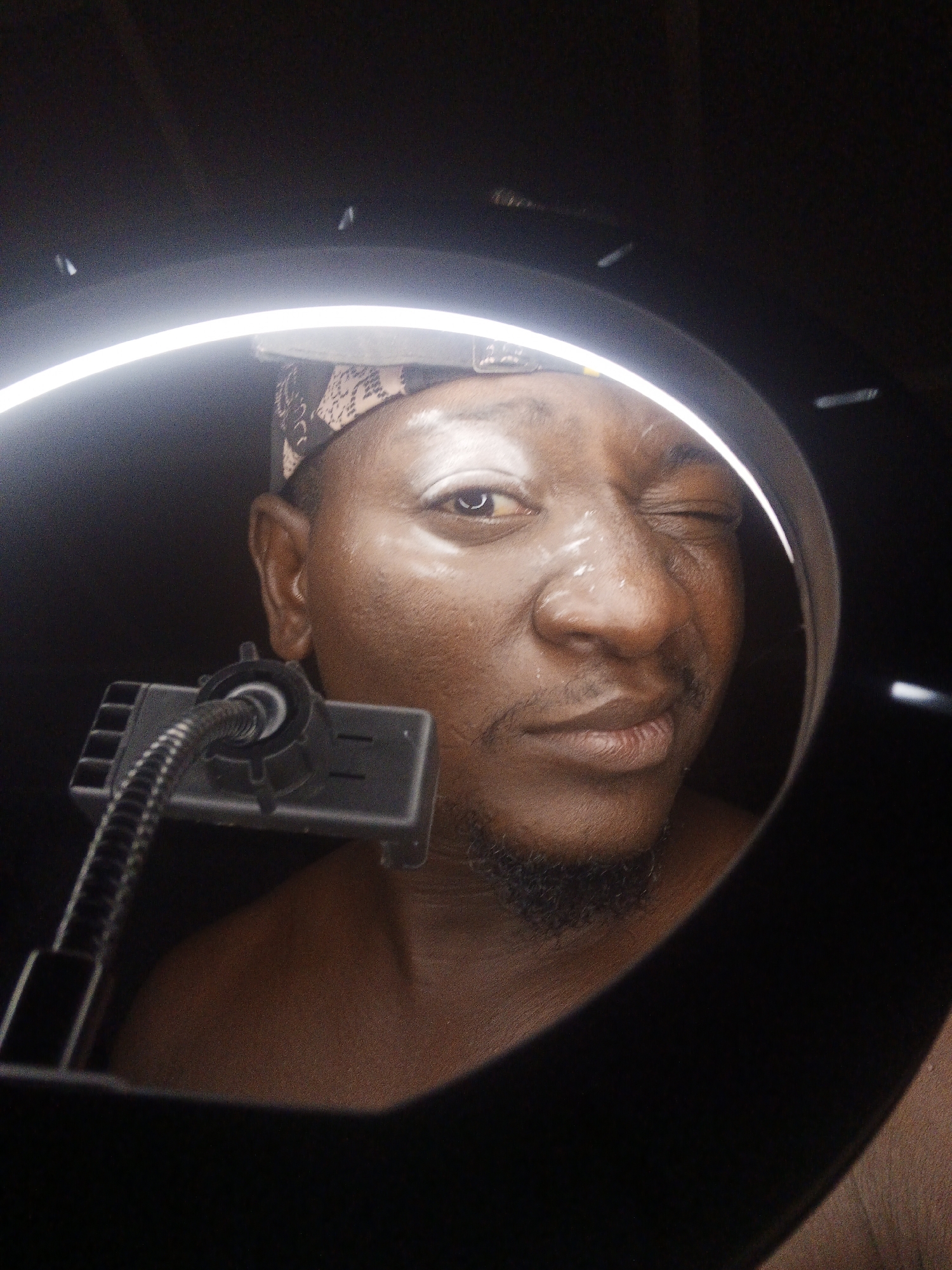 A close-up shot shows a man's face reflected in a ring light. The man is looking directly at the camera, winking one eye. His skin is dark, and he has a light-colored bandana on his head. The ring light creates a bright, circular frame around his face, with some light reflecting off his forehead and nose. A black, flexible mount with a device attached is visible in the foreground, obscuring part of his neck and chest. The background is dark and out of focus. The scene appears to be indoors, possibly in a studio or bedroom.