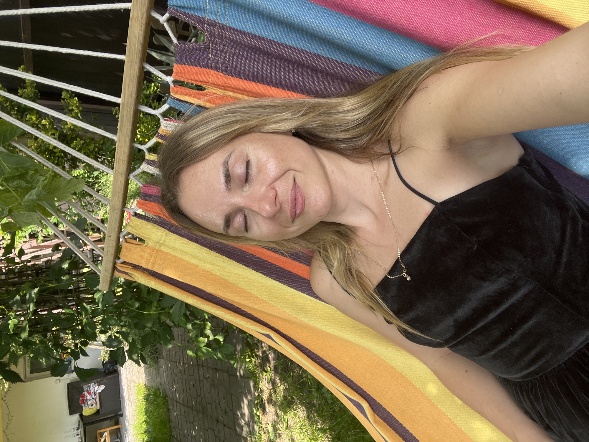 In a garden setting, a blonde woman with closed eyes is relaxing in a striped hammock. She is wearing a black dress and a delicate necklace. The hammock is suspended between two wooden beams, with ropes holding it in place. Lush green foliage surrounds the hammock, and a paved walkway is visible in the background. The overall scene suggests a tranquil moment outdoors.