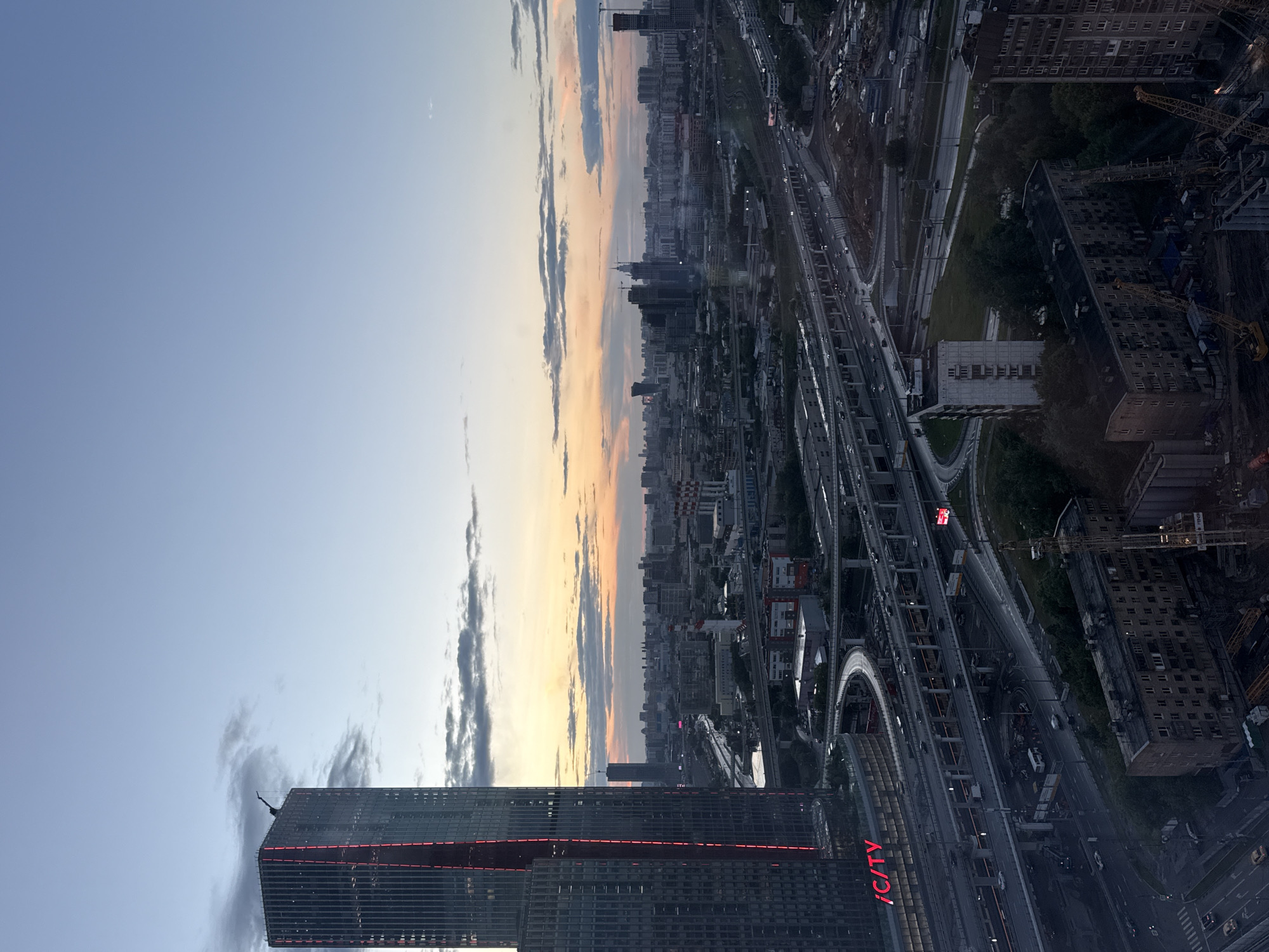This is a high-angle, wide shot of the cityscape of Khimki, Russia, during twilight. A prominent skyscraper, identified by the red "ICITY" signage, dominates the left side of the frame. The building features vertical red lighting accents. Surrounding the skyscraper is a complex network of elevated highways and overpasses, with traffic visible on them. In the mid-ground and background, a sprawling urban landscape unfolds, characterized by numerous buildings of varying heights, including residential blocks and other high-rises. The sky displays a gradient of colors from light blue at the top to hues of orange and pink near the horizon, indicative of sunset or sunrise. Scattered clouds add texture to the sky. The overall scene depicts a densely populated urban environment with extensive infrastructure.