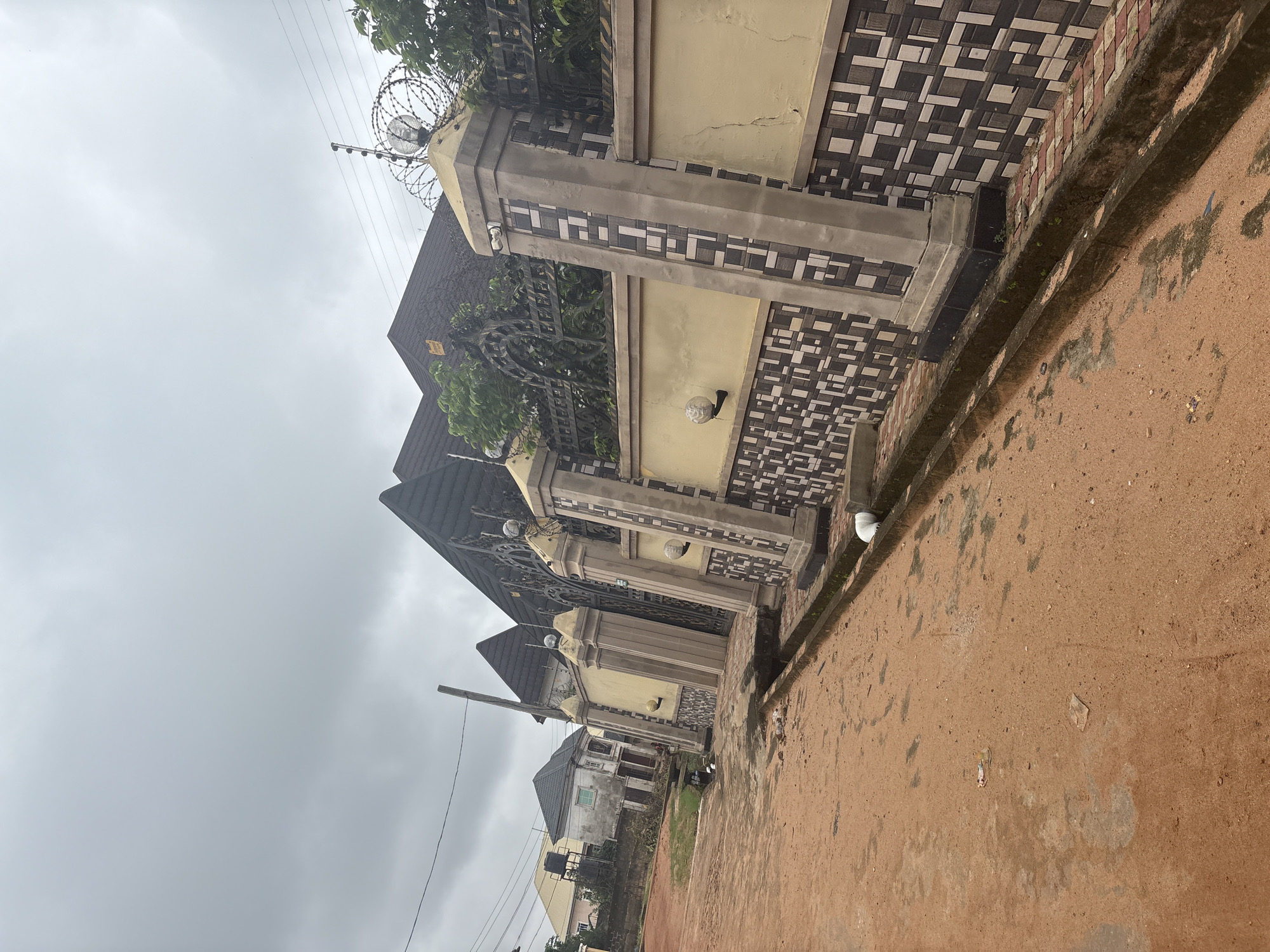 This image shows a residential street in Obosi, Nigeria, under an overcast sky. The foreground is a dirt road with some scattered litter. To the right is a property with a tall, decorative wall and a wrought-iron gate. The wall is capped with concrete pillars that have a mosaic tile pattern. Barbed wire and a security light are visible on top of one of the pillars. Beyond the wall, parts of the building's dark-tiled roof and some greenery are visible. In the background, other houses with similarly styled roofs and walls can be seen, along with a water tank on one of the buildings. The overall impression is of a well-maintained, possibly affluent, residential area.