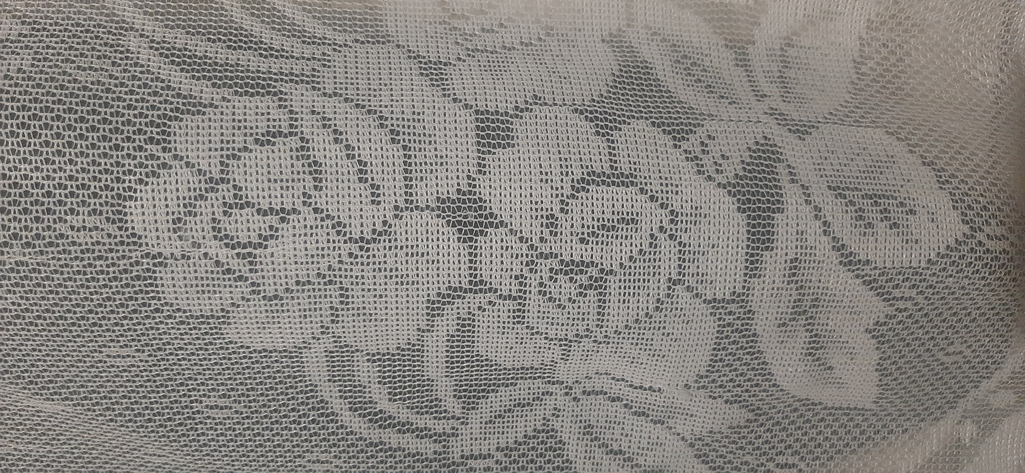 A white lace curtain is shown against a dark background. The lace has a floral pattern. The image is a close-up, capturing the texture and intricate design of the fabric. There are no people or any other objects visible in the frame. The setting is indoors, with the curtain likely covering a window. The lighting is diffused, suggesting an indoor environment with soft light filtering through the curtain. No specific location in Yessentukskaya, Russia, can be determined from the image alone.