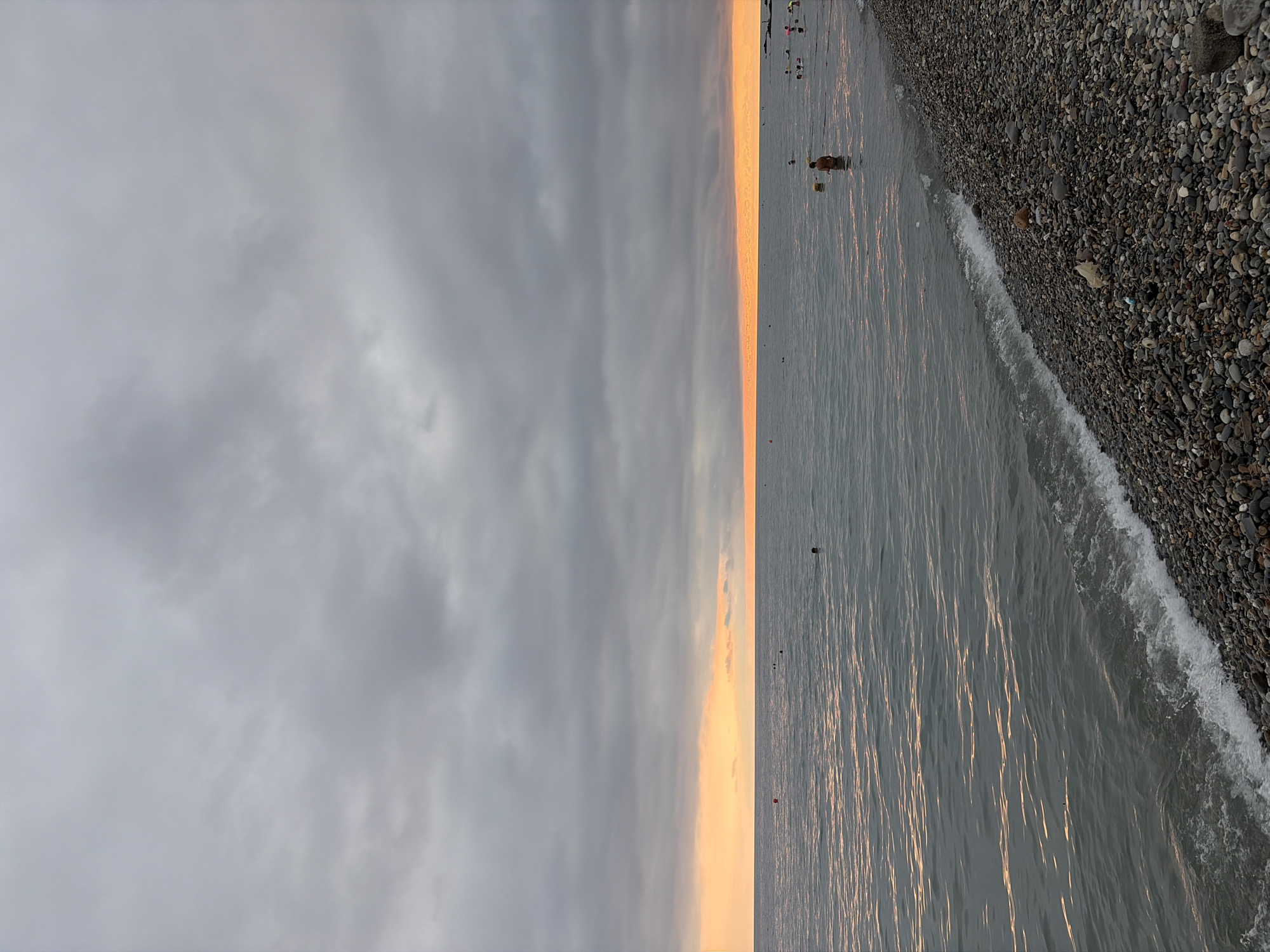 The image depicts a pebble beach and the sea at sunset in Sochi, Russia. The sky is overcast with dark grey clouds, but a strip of vibrant orange and yellow hues is visible along the horizon where the sun is setting. The sea is calm, with gentle waves lapping onto the shore. Several people are in the water, some swimming and others wading. One person is standing in the water near the shore, facing away from the camera. A few buoys are scattered in the sea. The pebble beach is covered with an array of small, smooth stones of various colors. The overall atmosphere is tranquil and serene, with the fading light casting a warm glow on the scene.