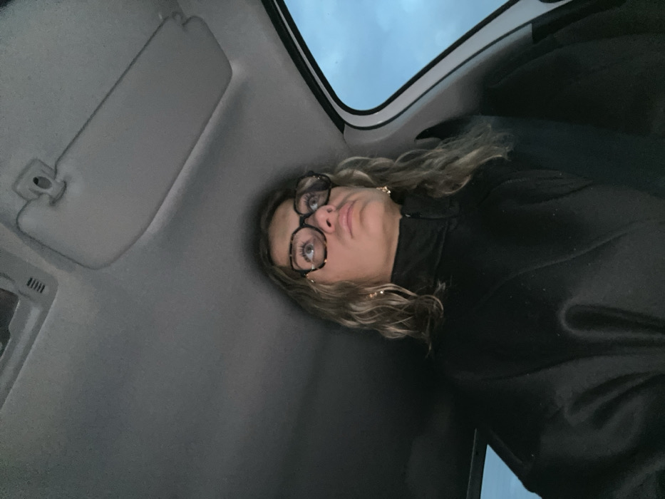 A person with glasses and curly hair is positioned in a vehicle. The camera angle is low, capturing the person from below. They are wearing a dark-colored jacket. The vehicle's interior includes a sun visor and a view of the sky through the roof. The person's expression appears somewhat quizzical or pensive.