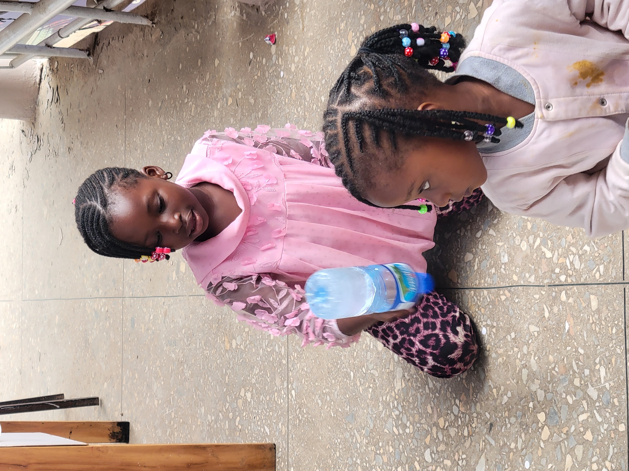 Two young Black girls are on the floor, likely indoors given the flat, tiled surface. The girl on the left wears a pink dress with floral details and leopard-print slippers. She holds a bottle of water. The girl on the right wears a light pink jacket over a grey shirt. Both girls have braided hair adorned with colorful beads. The location is presumed to be Jos, Nigeria, based on the prompt's context.