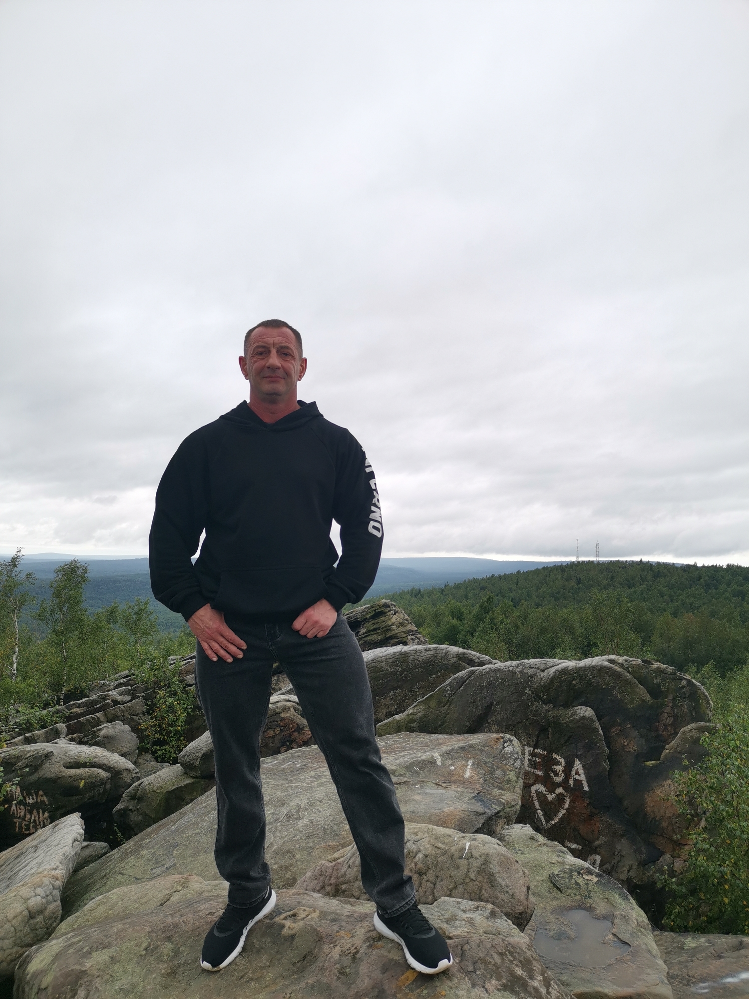 A man stands on a rocky outcrop in the city of Gubakha, Russia, with a forest-covered valley and a cloudy sky in the background. The man is wearing a black hooded sweatshirt with white lettering on the sleeve, dark wash jeans, and black athletic shoes. He is looking directly at the camera with his hands in his pockets. The rocks in the foreground are large and weathered, with some graffiti visible on them, including a heart shape and some Cyrillic lettering. The background shows rolling hills covered in dense green trees, with two radio towers visible on a distant hill. The overall lighting suggests an overcast day.