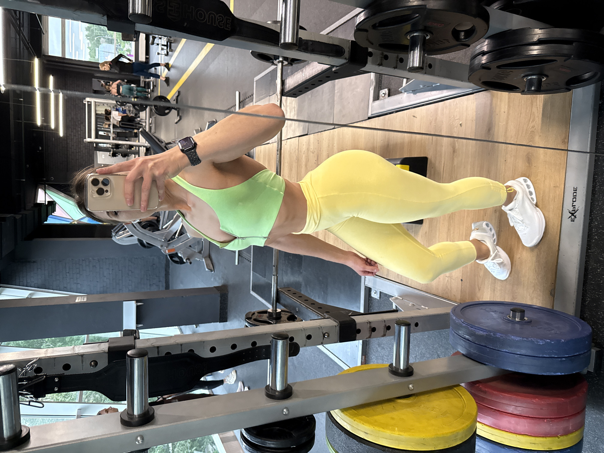 A woman in yellow leggings and a green top takes a selfie in a gym. She holds a phone in her left hand, displaying a watch on her wrist. The gym equipment includes weight racks with weight plates, and workout machines. Other individuals are visible in the background. The gym appears to be located in Saint Petersburg, Russia.