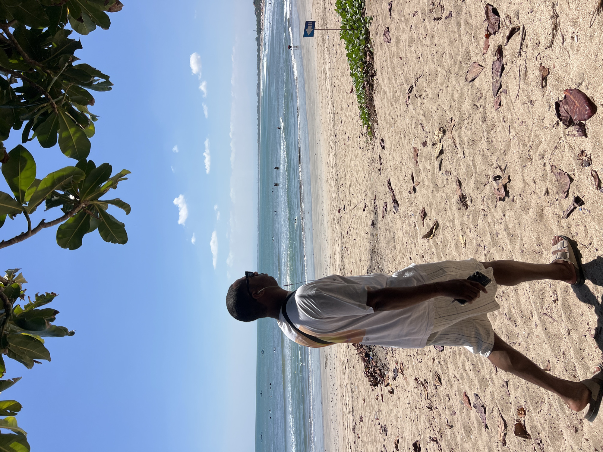 In Kuta, Indonesia, a person walks along a sandy beach with the ocean in the background. The person is wearing a white t-shirt with a colorful graphic on the back, light-colored striped shorts, sunglasses, and white earphones. They are holding a smartphone in their right hand. The beach is covered in sand and scattered with fallen leaves. In the distance, the ocean has gentle waves, and several people can be seen in the water, some appearing to be surfing or wading. A blue directional sign with a white arrow is planted in the sand to the right of the frame. The sky is a clear blue with a few wispy clouds, and the sun casts shadows on the sand. Lush green foliage hangs from branches in the upper portion of the image, framing the scene.